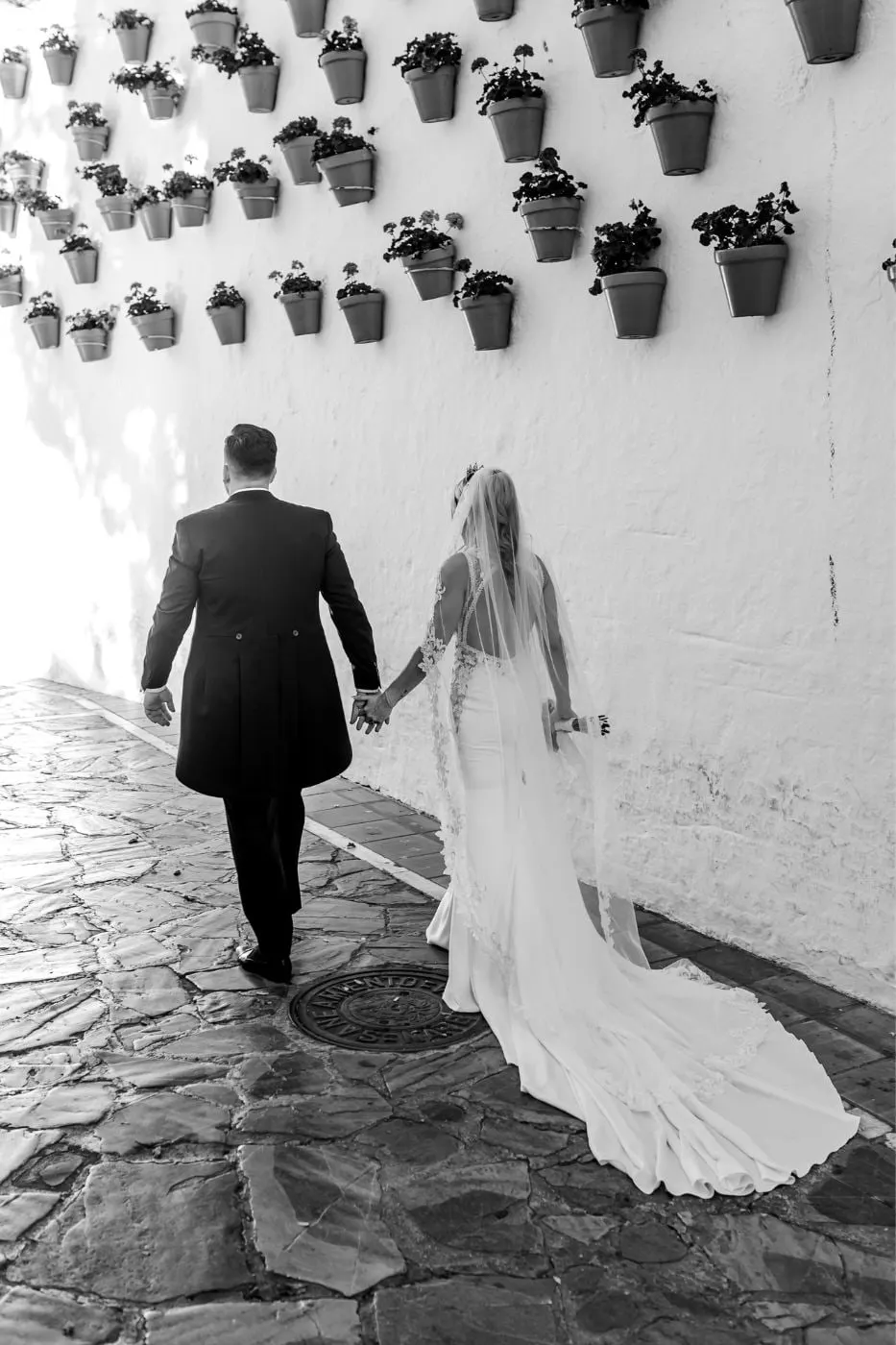 Black and white of the bride and groom walking hand in hand up a whitewashed street lined with flowerpots, Costa del Sol