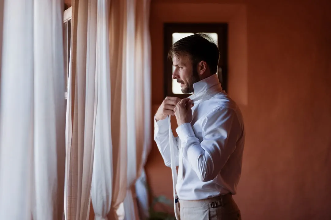Groom buttoning his collar by a curtained window against a terracotta wall, Costa del Sol