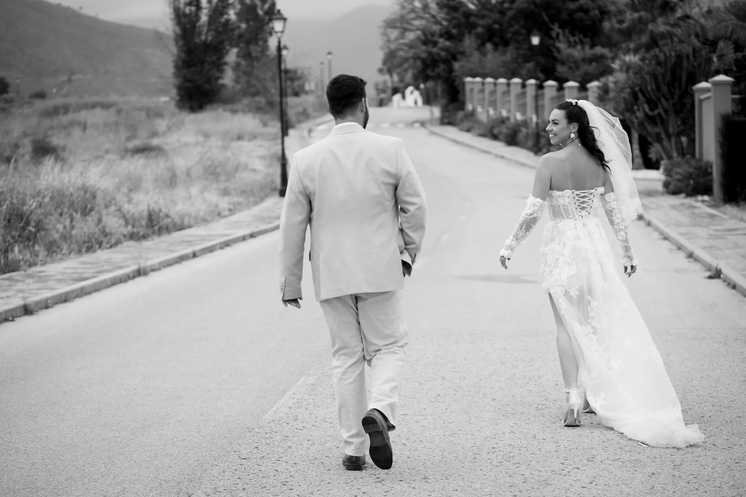 Black and white of the bride and groom walking up the road at Hacienda San José, the bride looking back with a smile, Mijas