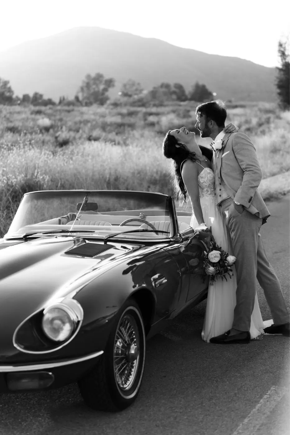 Black and white of the couple kissing beside a classic open-top car, hills behind, Mijas