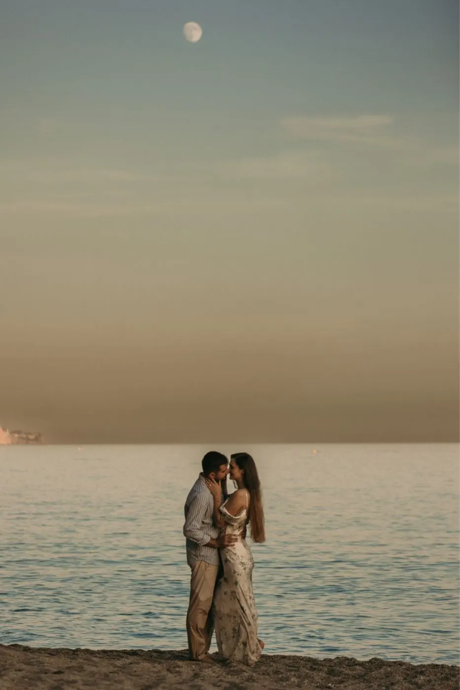Bride and groom embracing at the water's edge at sunset, the moon above the sea, Nerja