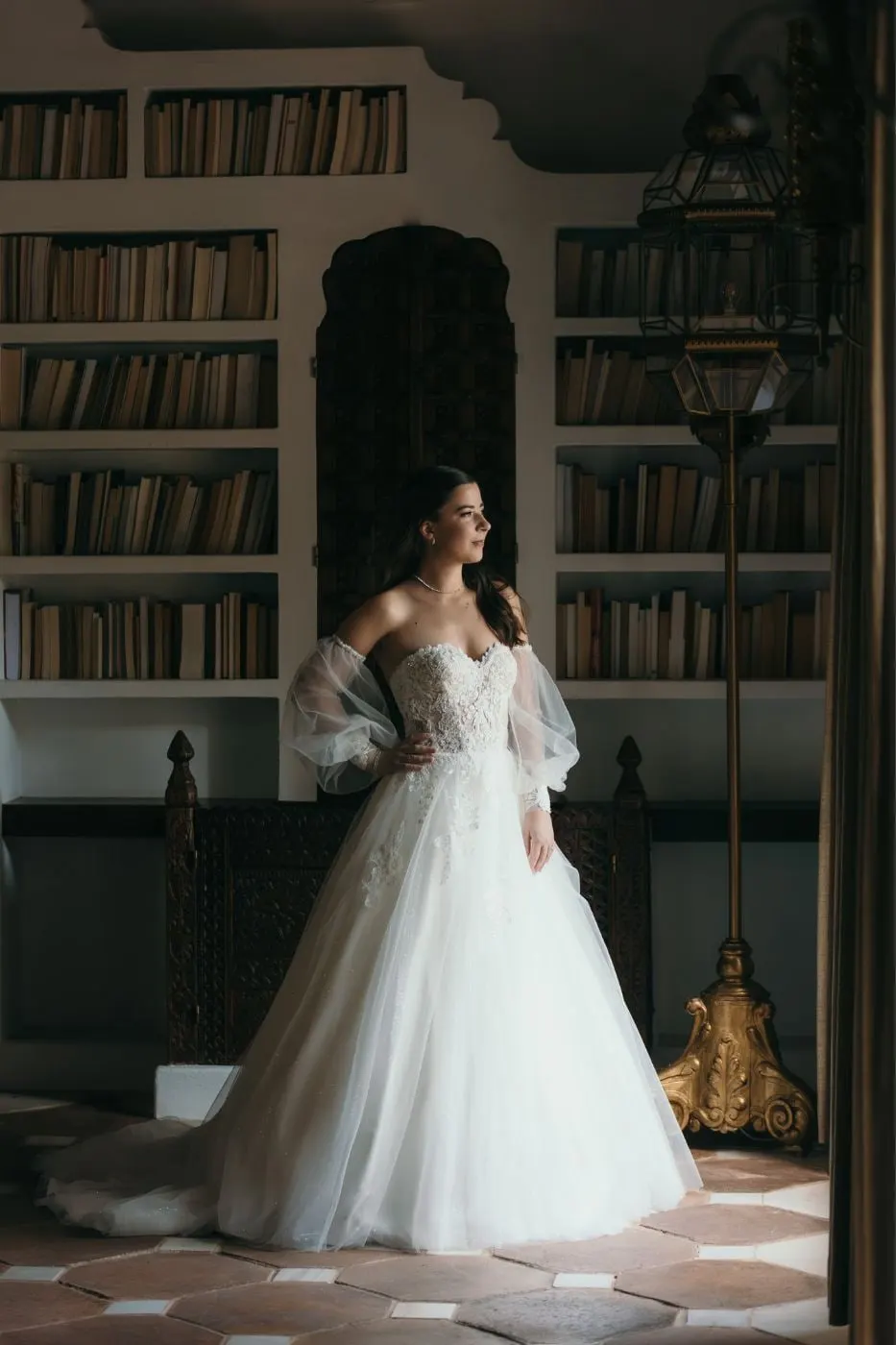 Bride in a strapless gown standing by tall bookshelves in a library, Cortijo San Francisco, Estepona