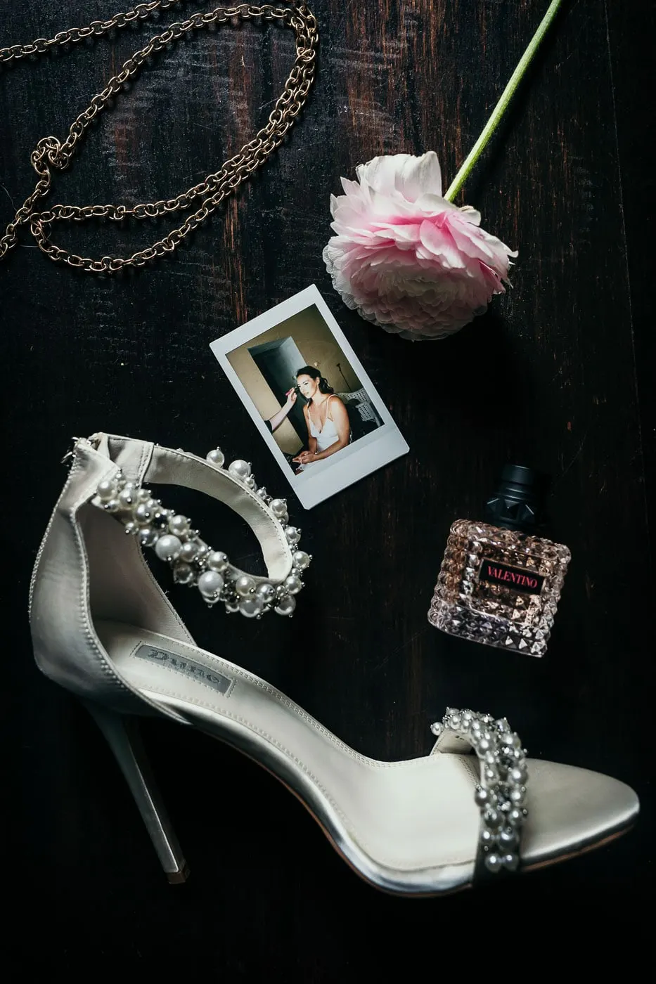 Pearl-studded heels, a pink ranunculus, perfume and a Polaroid laid on dark wood, Hacienda San José, Mijas