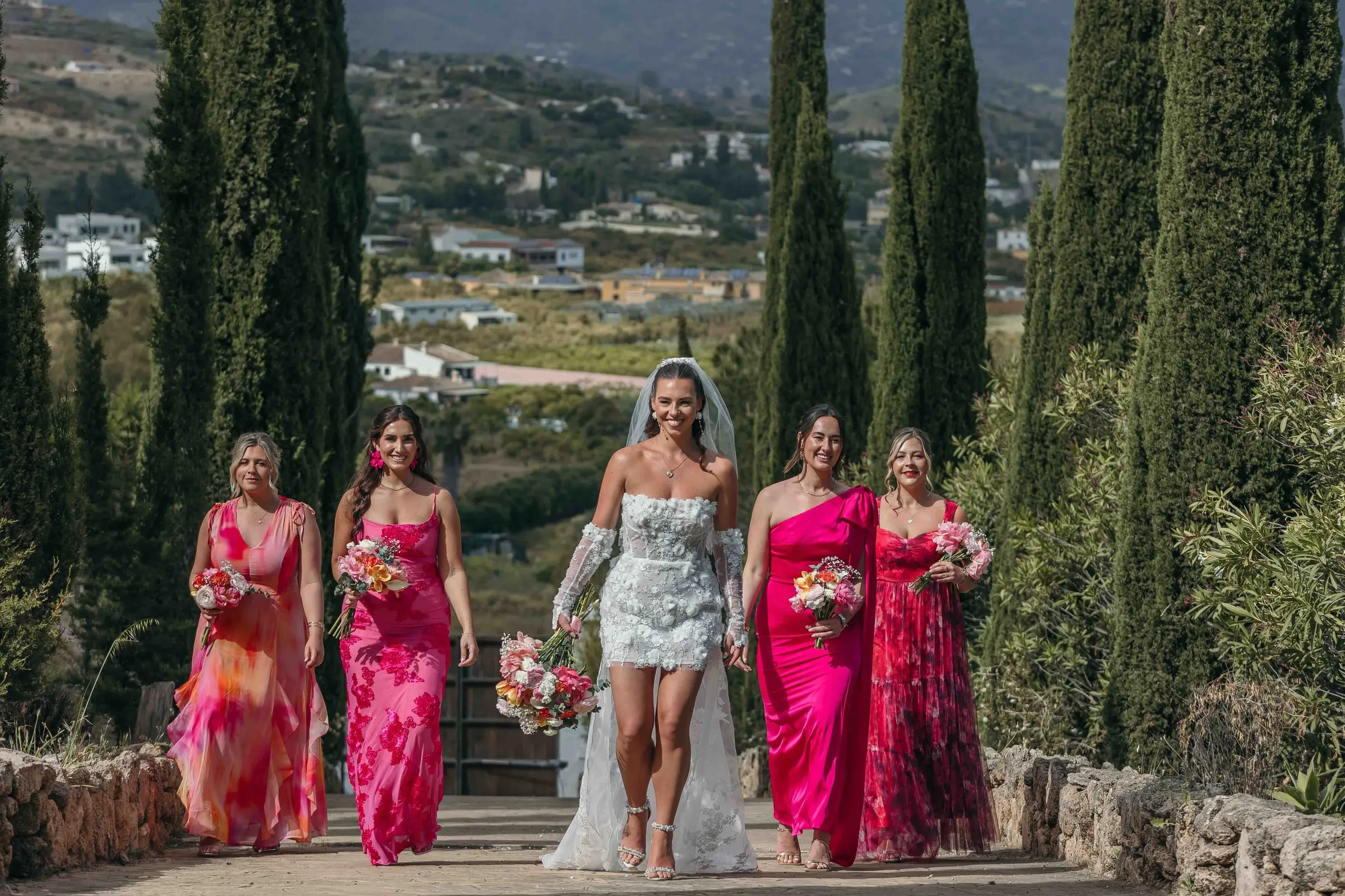 Bride walking with her bridesmaids in pink between cypress trees, destination wedding in Málaga