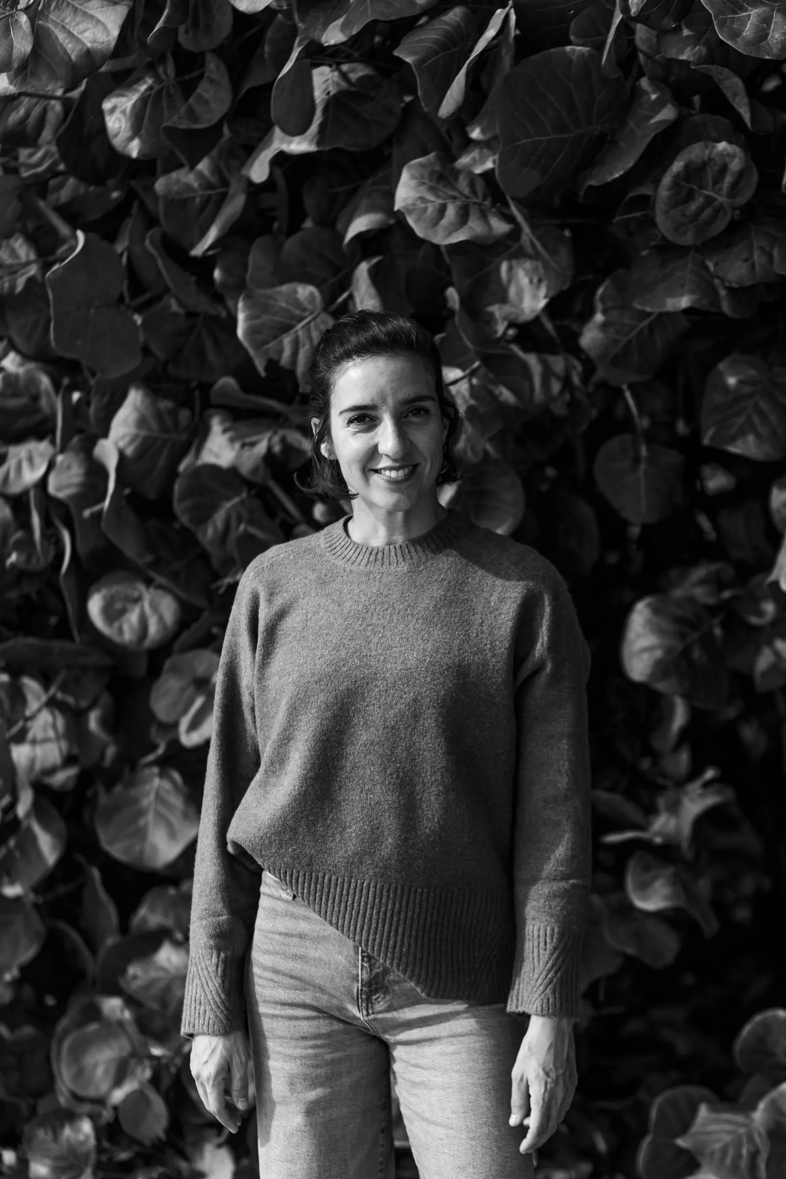 Black and white portrait of Tamara, Picart's videographer, smiling against a wall of leaves, Estepona