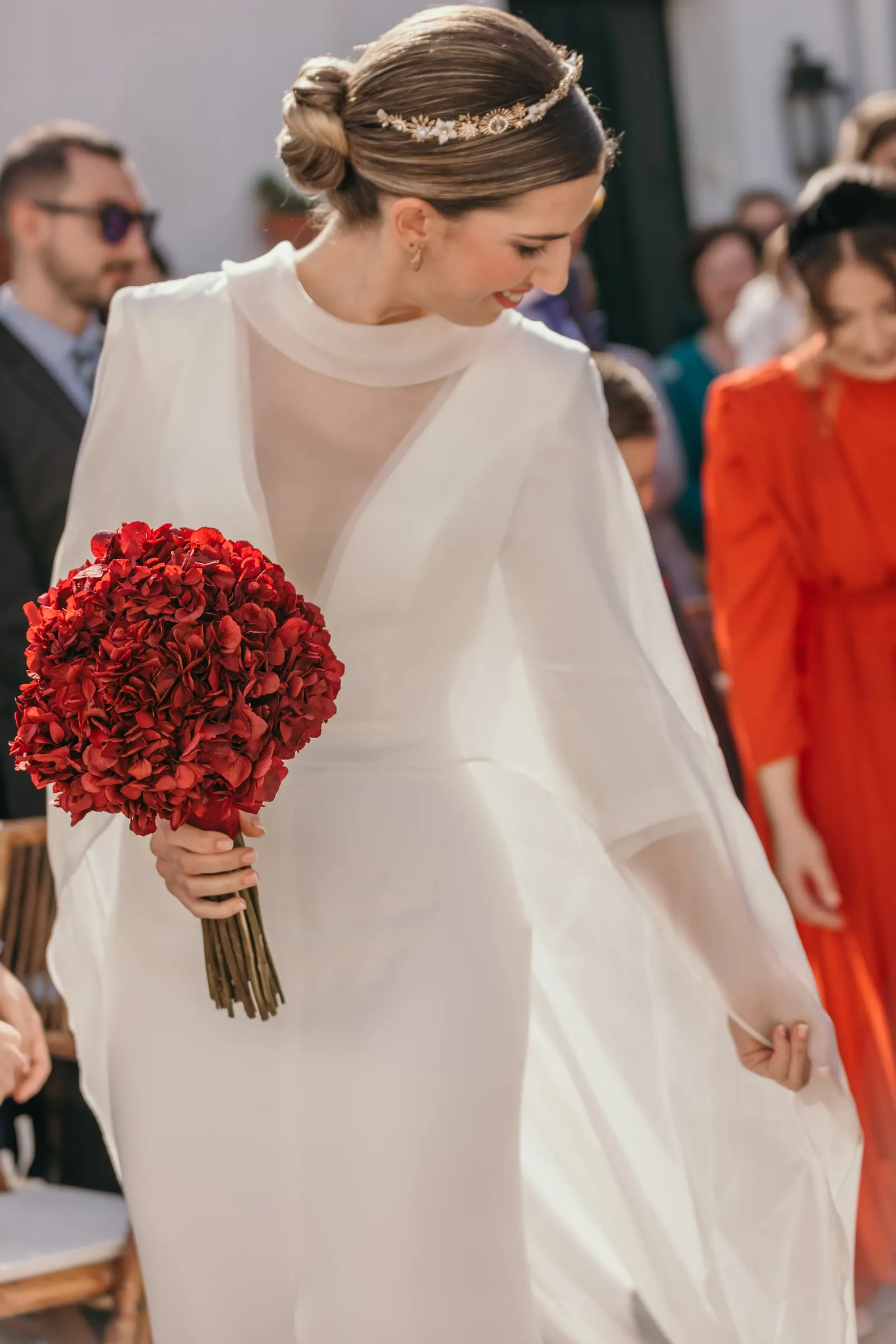 Bride in a high-neck cape dress with a deep red rose bouquet arranging her train before the ceremony