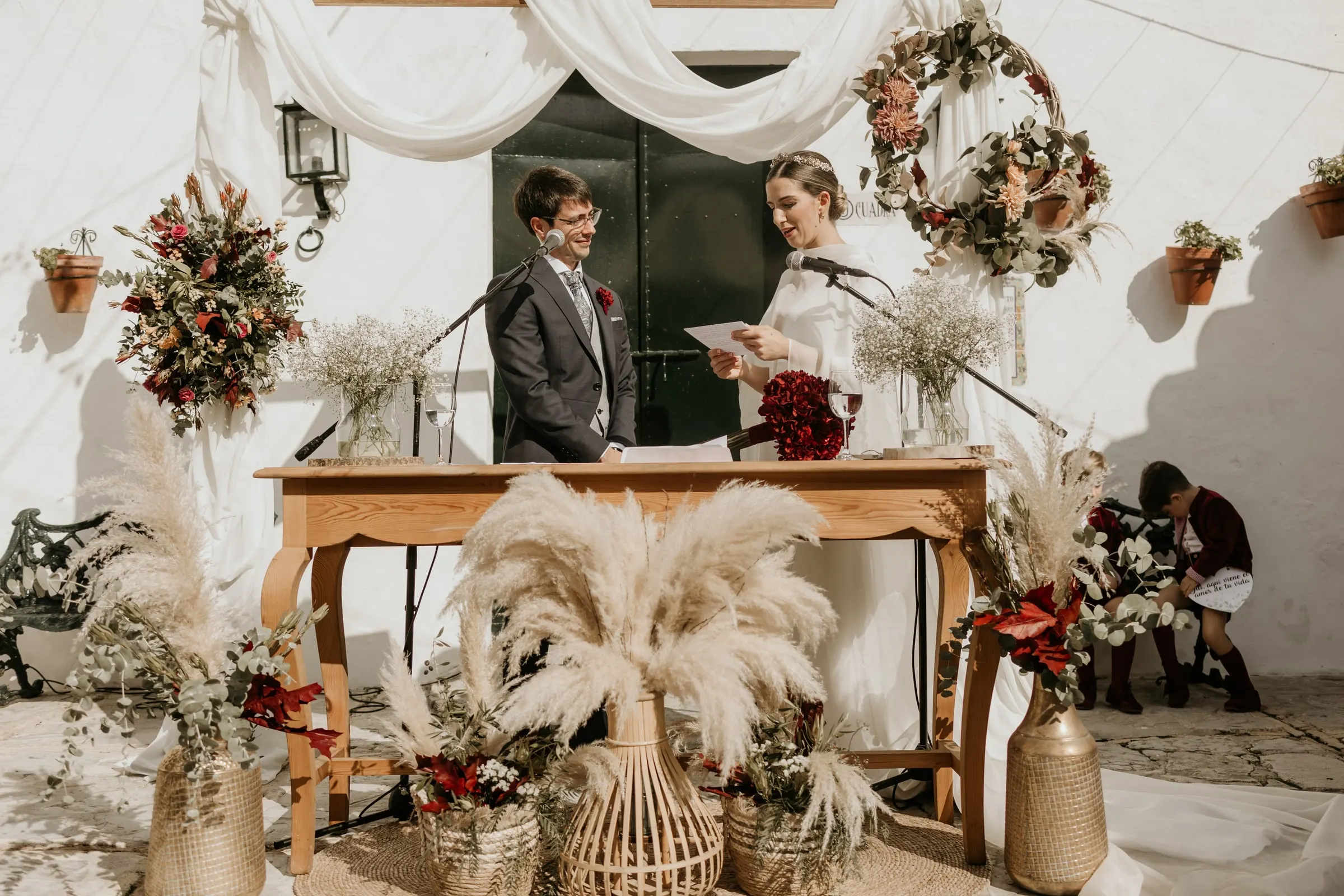 Couple at the ceremony table under white drapes, dried flower wreaths and lanterns in the cortijo courtyard