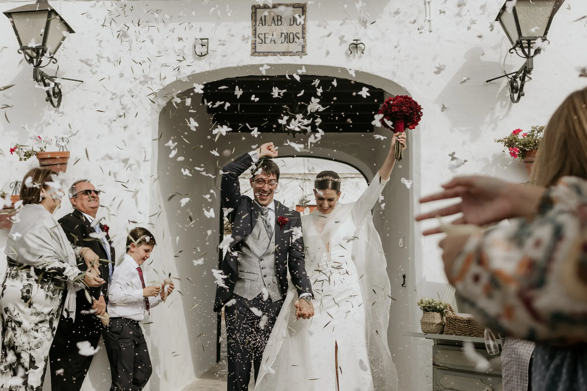 Newlyweds leaving through the cortijo archway under a shower of paper petals and an 'Alabado sea Dios' tile