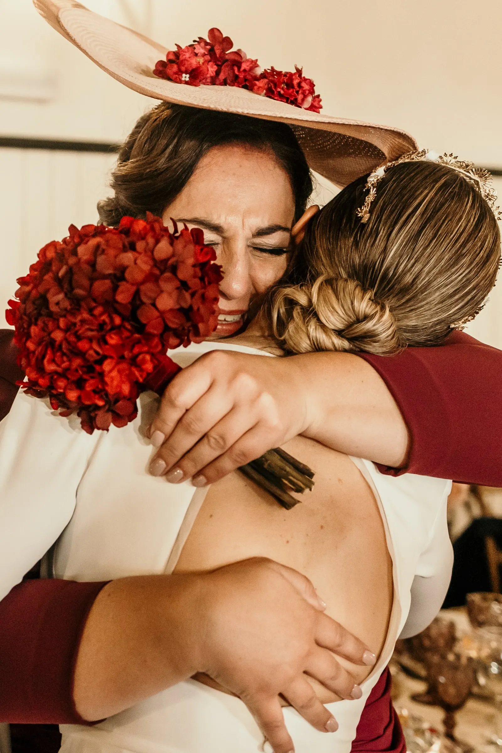 Bride hugging a guest in a wide-brimmed hat with her red bouquet in hand