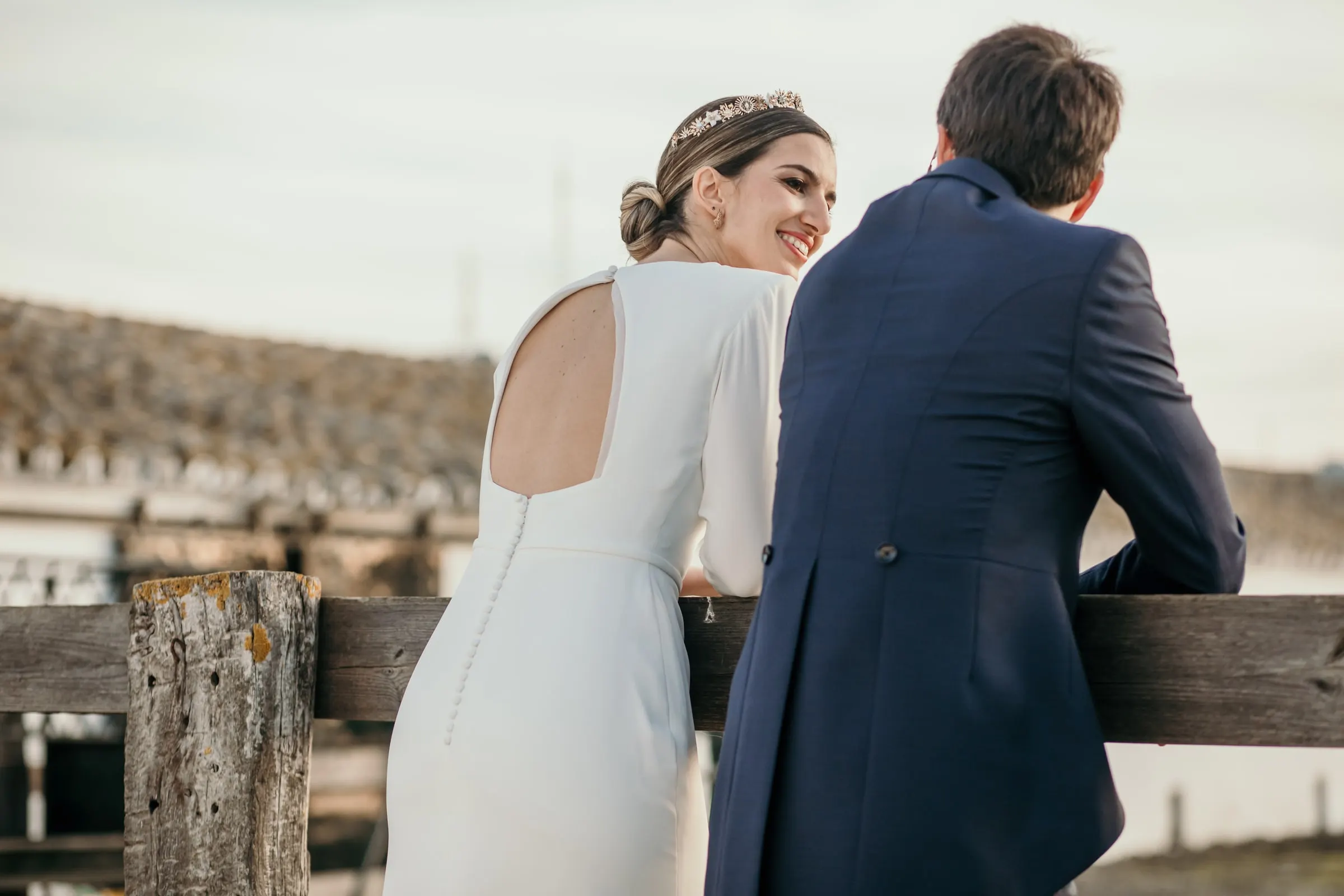 Bride smiling over her shoulder in an open-back dress beside the groom at the fence