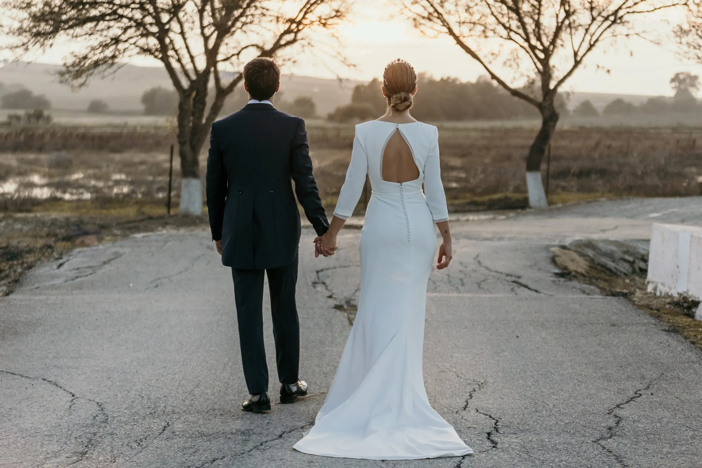 Newlyweds walking hand in hand down a country lane between bare trees at sunset