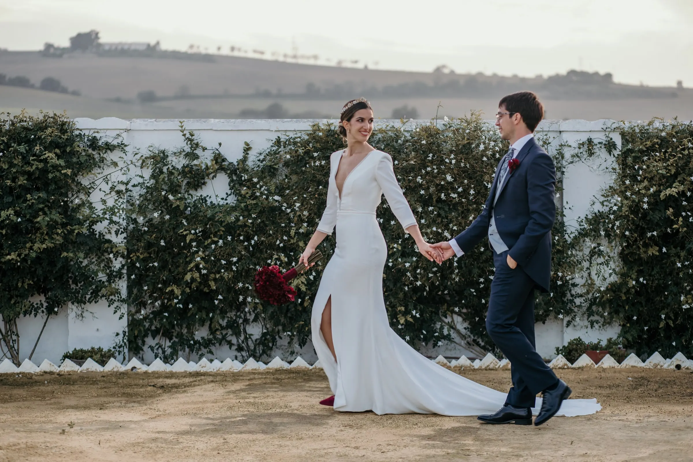 Bride leading the groom along a white wall covered in climbing plants with rolling hills behind