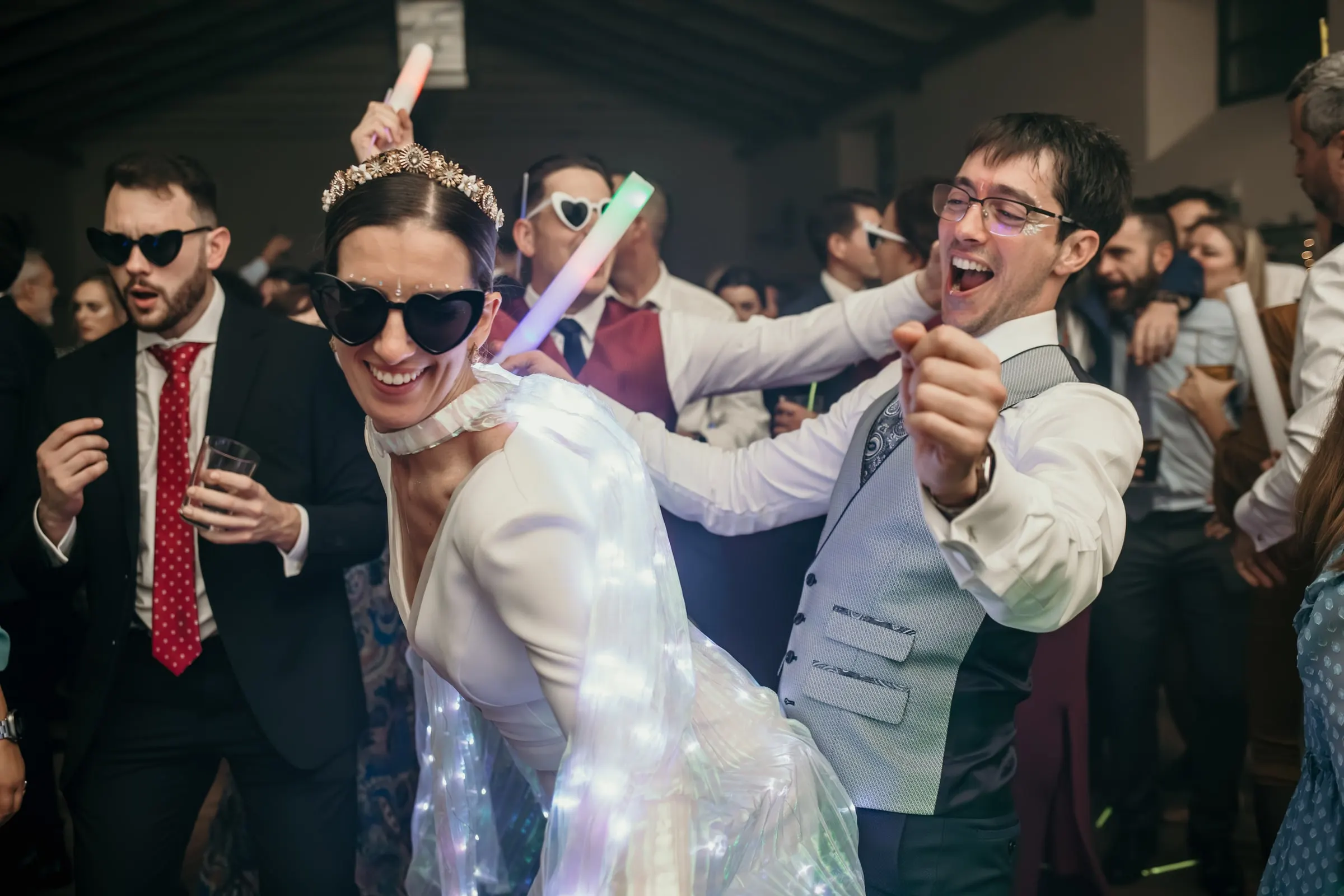 Bride in heart-shaped sunglasses dancing with the groom and friends late at night
