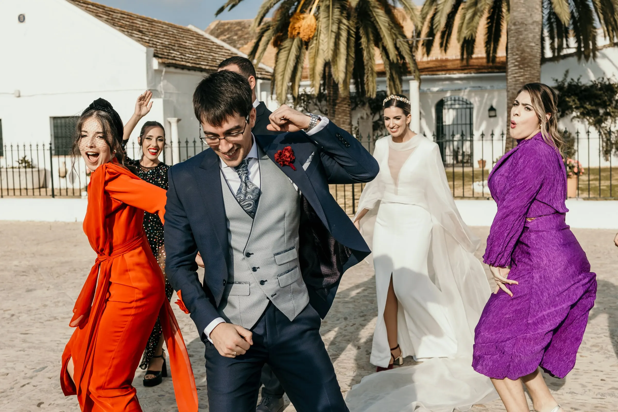 Groom dancing with friends in orange and purple dresses in the sunny yard of the Andalusian cortijo, bride behind