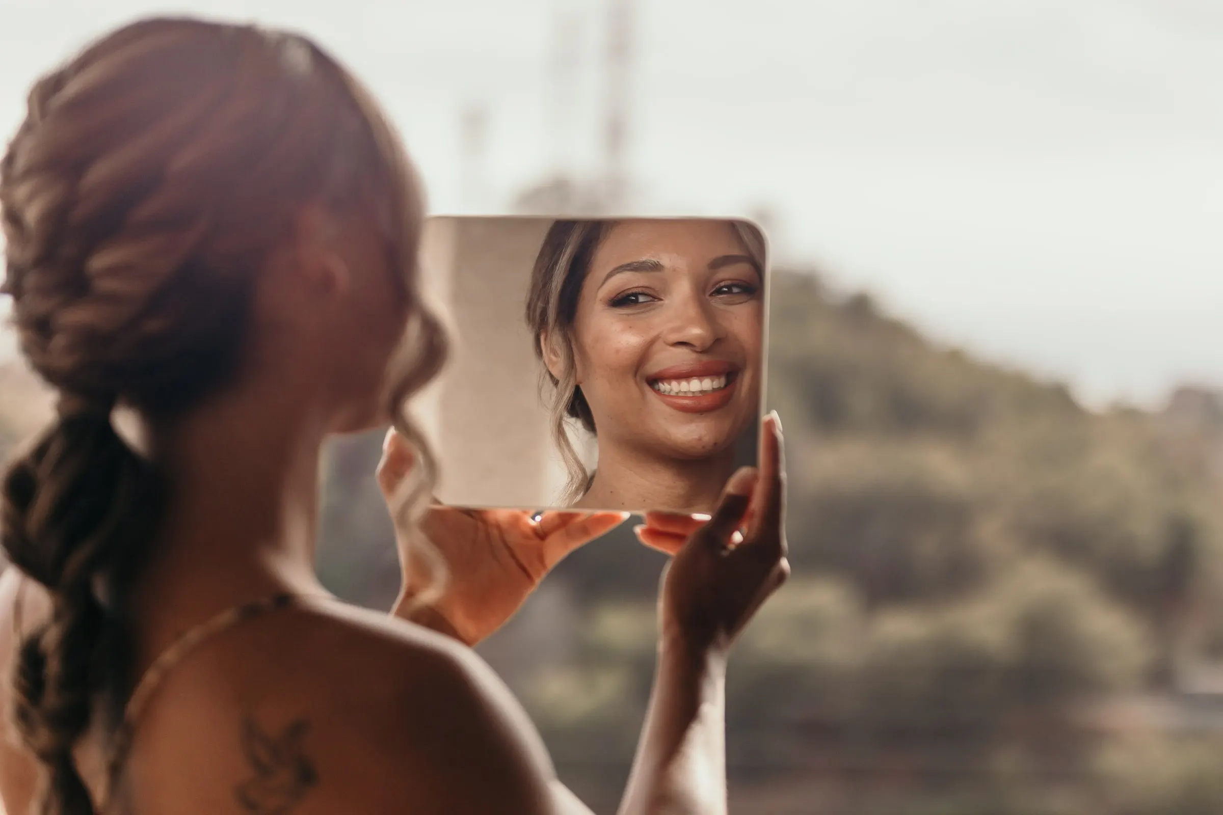 Bride with a braided ponytail smiling into a hand mirror against the green hills of the Costa del Sol