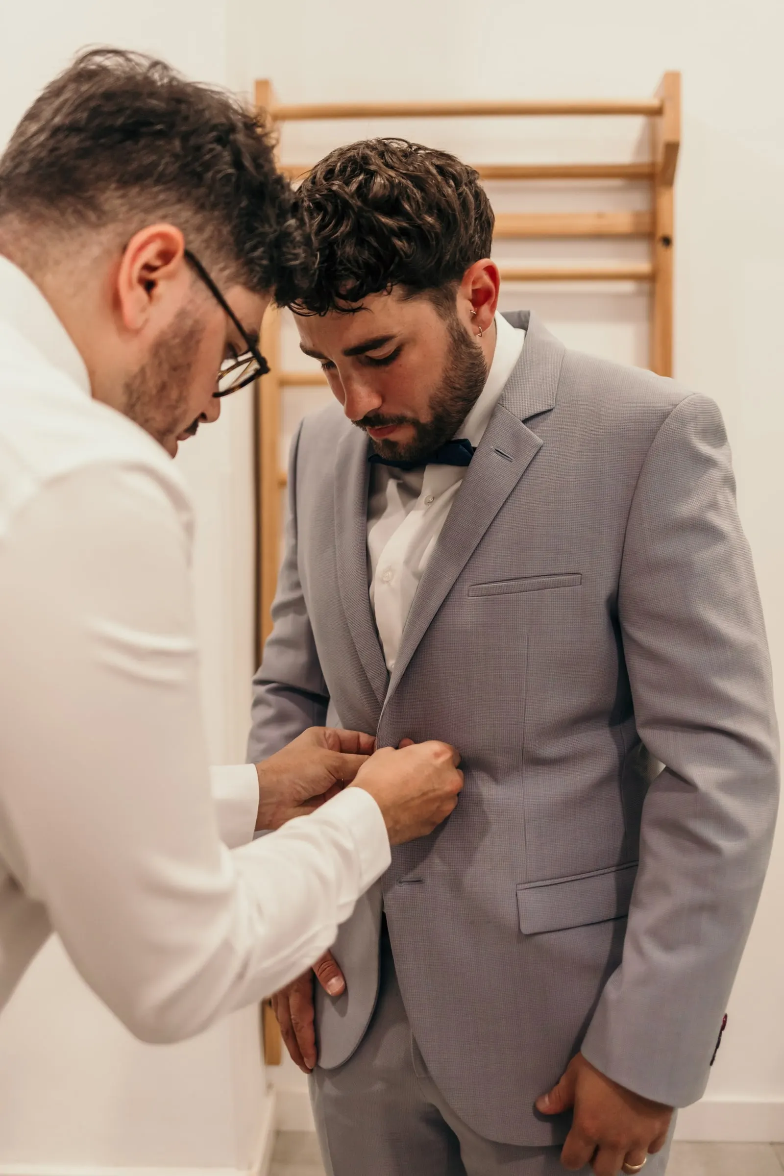 Friend buttoning the groom's pale grey jacket, foreheads almost touching