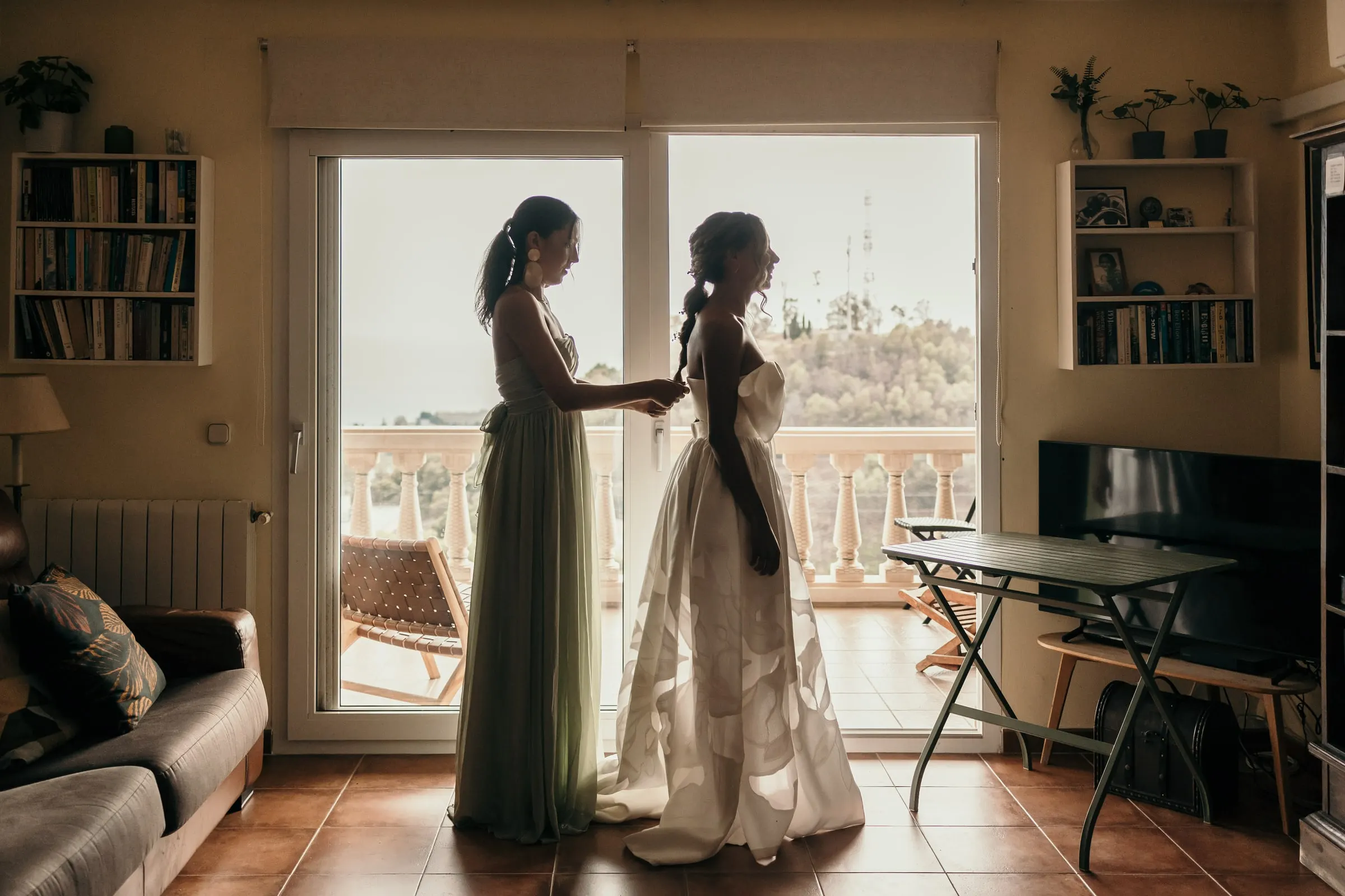Bridesmaid in sage green helping the bride into her dress by the balcony doors of a villa