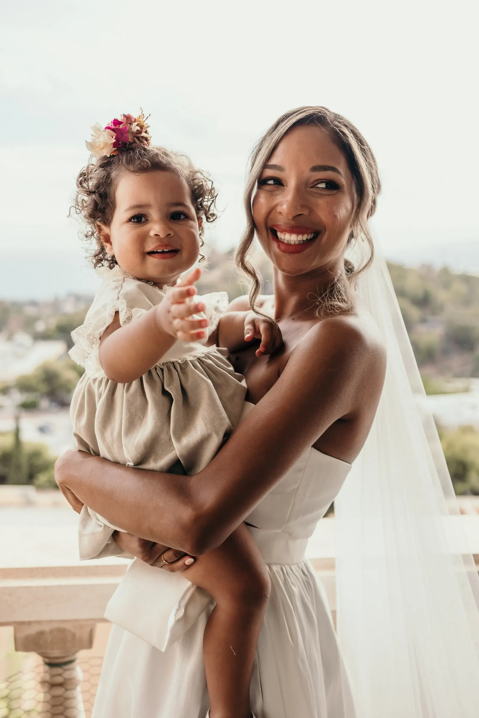 Bride in her veil holding a laughing toddler with flowers in her hair on the balcony