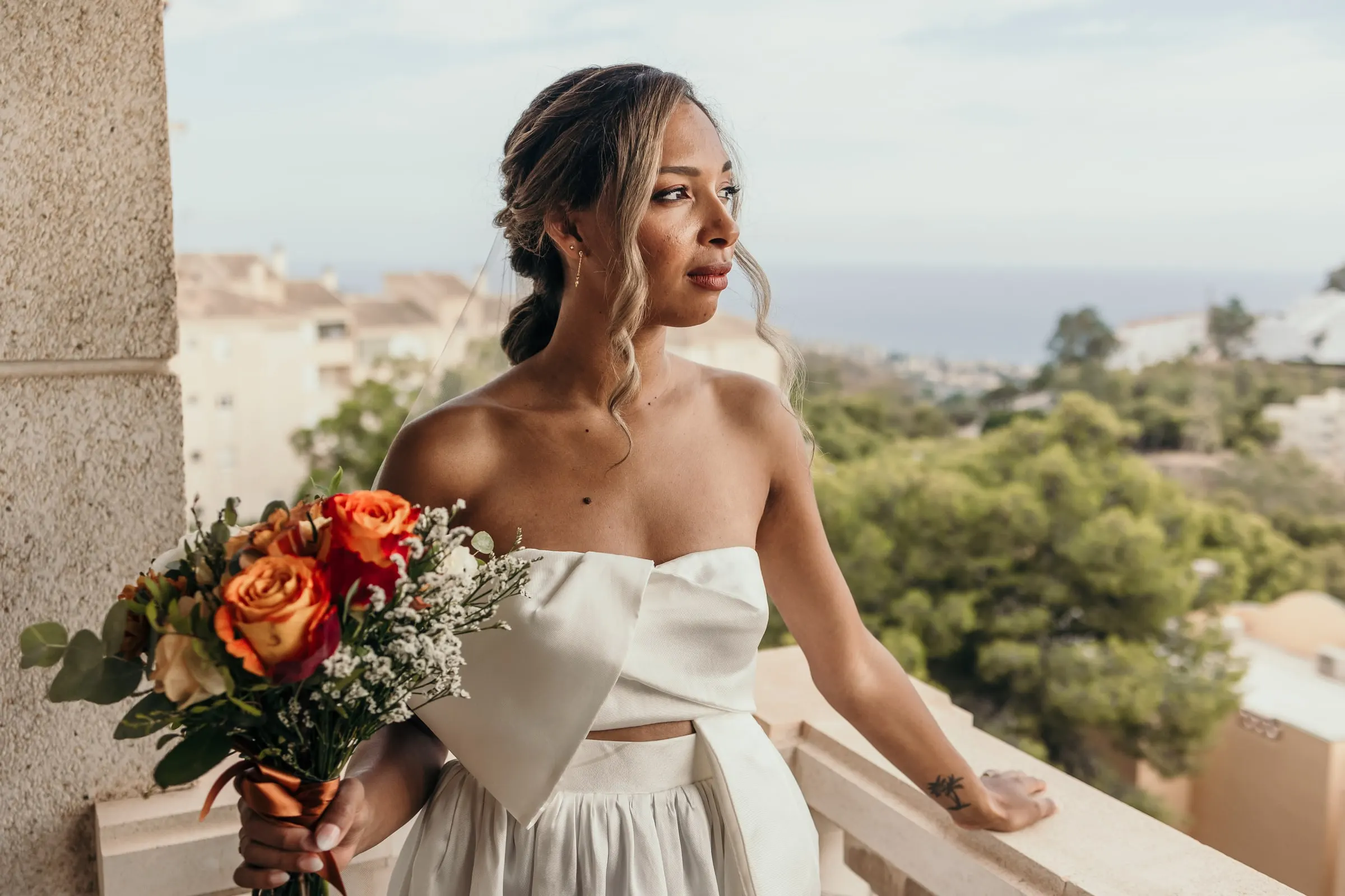 Bride holding her orange rose bouquet on a balcony with the Mediterranean on the horizon