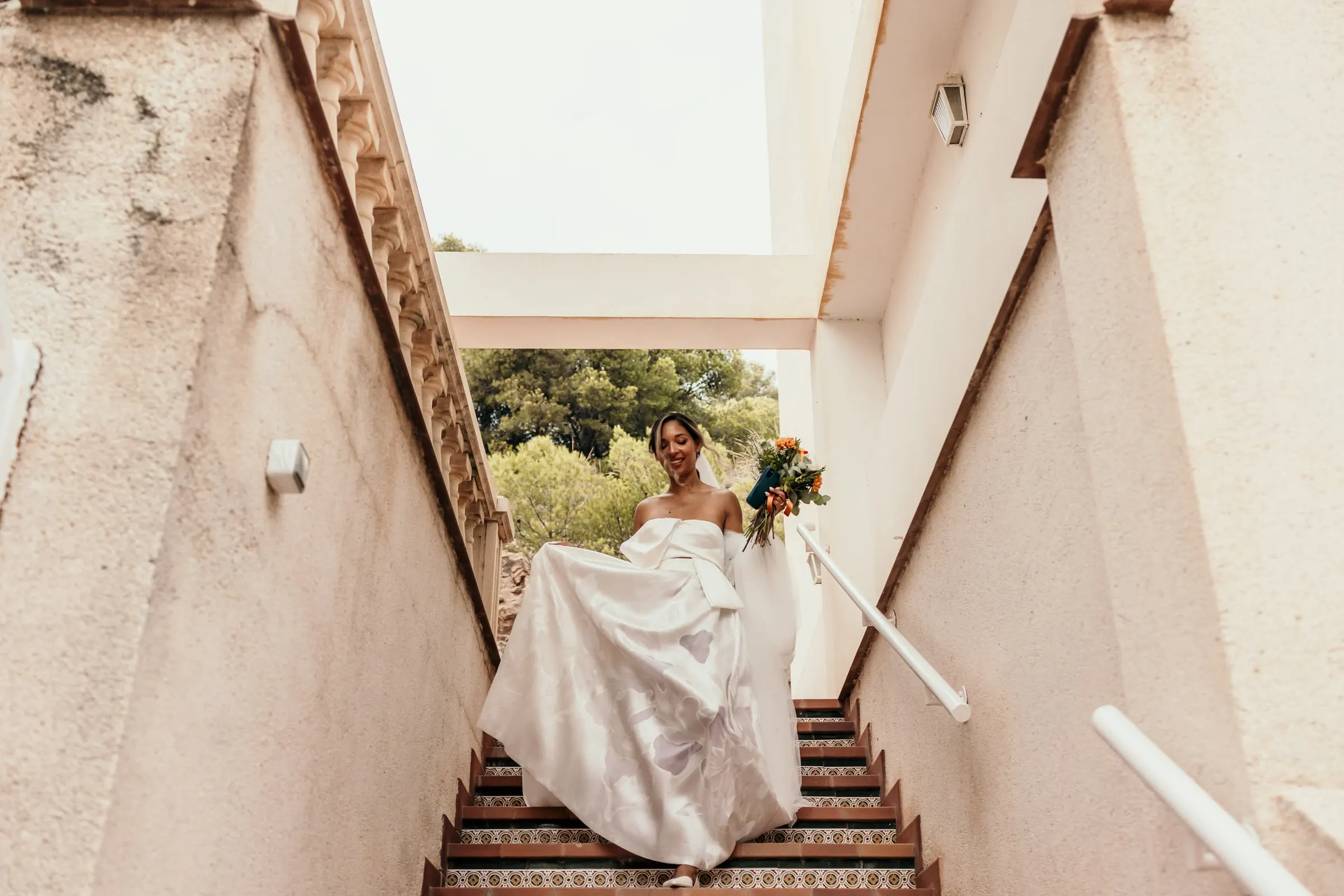 Bride lifting her skirt as she walks down an outdoor staircase between white walls