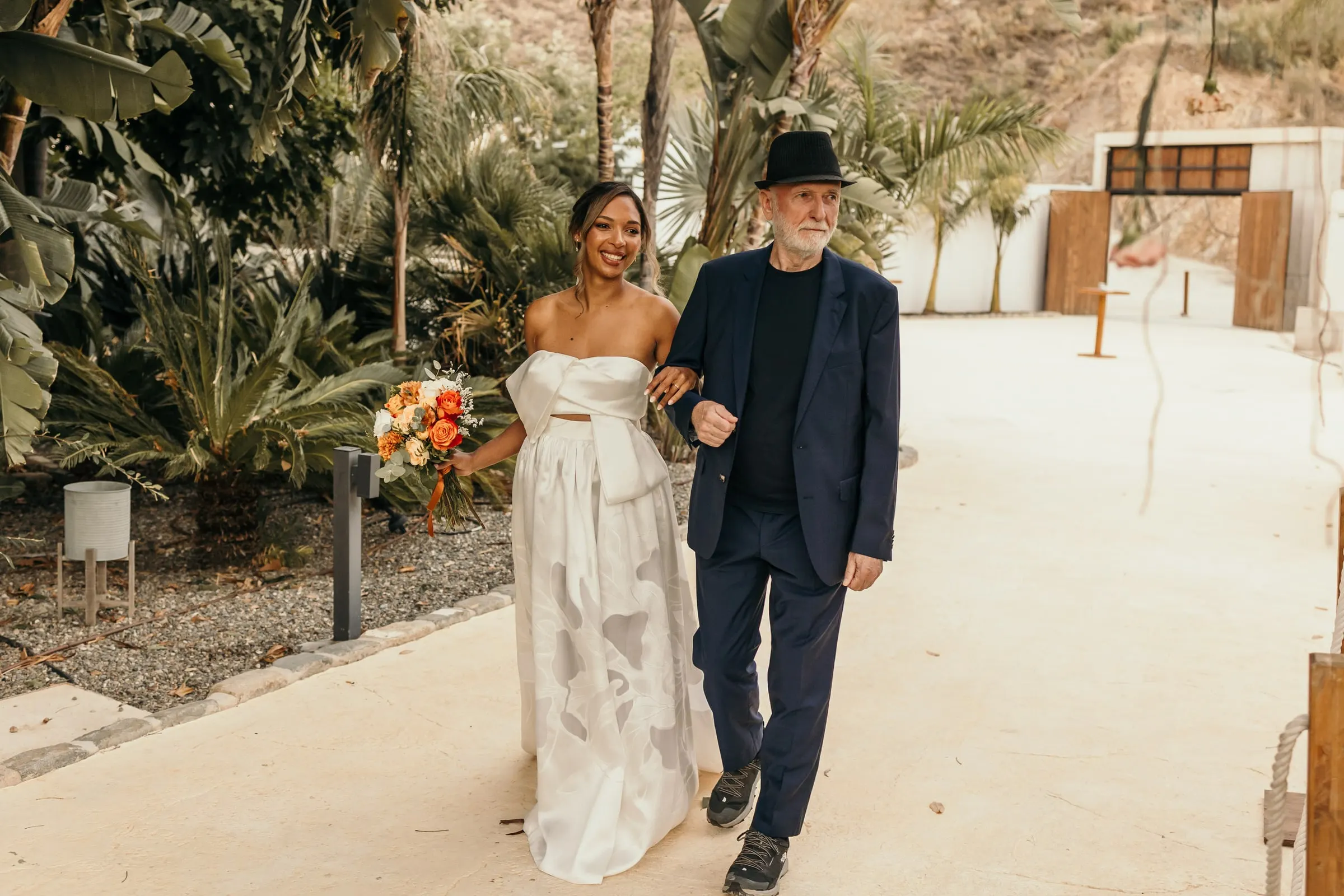 Bride arriving arm in arm with the man in the black hat through a tropical garden of palms