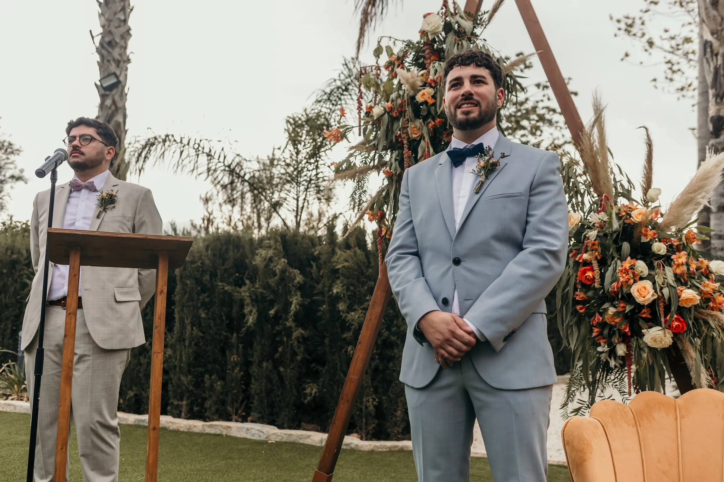 Groom in a pale blue suit and navy bow tie waiting by the floral arch for the outdoor ceremony