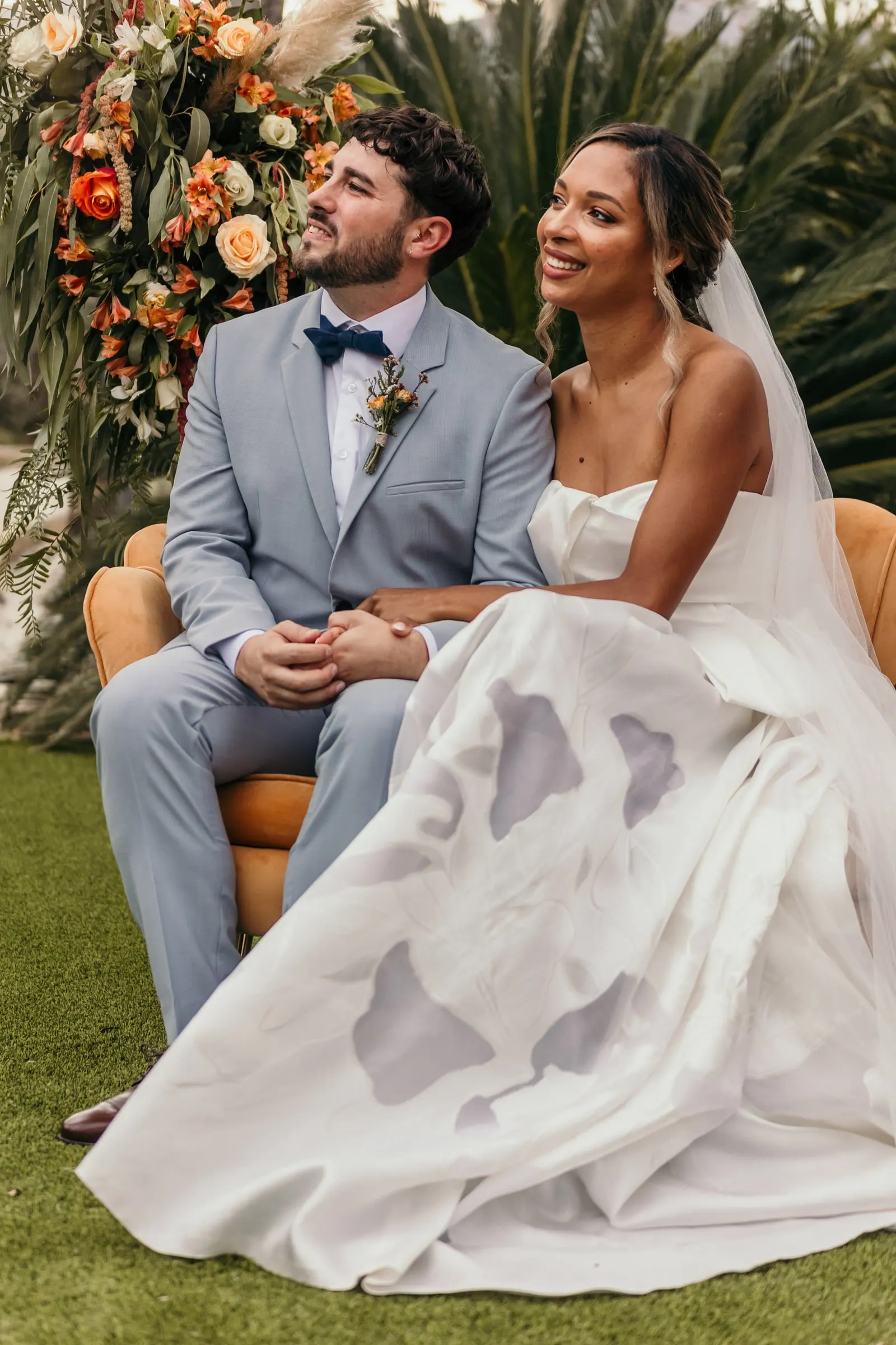 Bride and groom laughing on velvet armchairs beside the orange flower arrangement