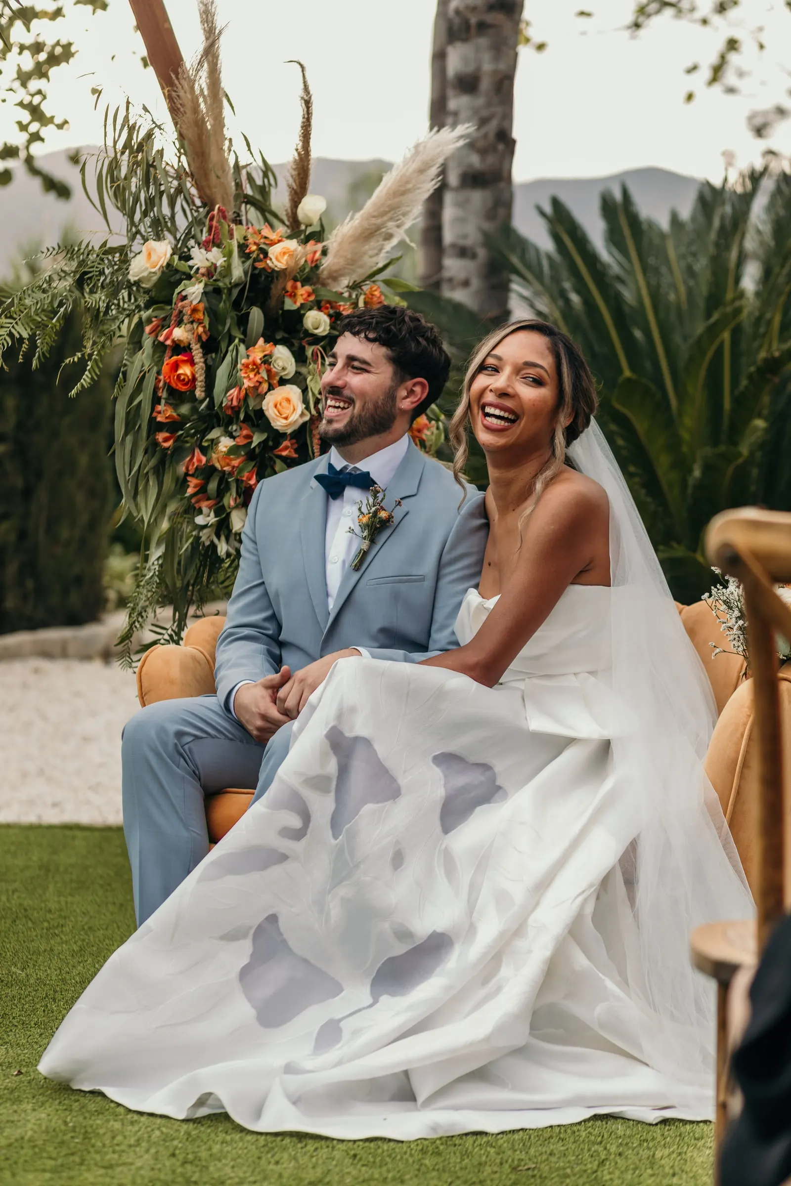 Bride and groom laughing at the ceremony under the pampas grass and orange roses