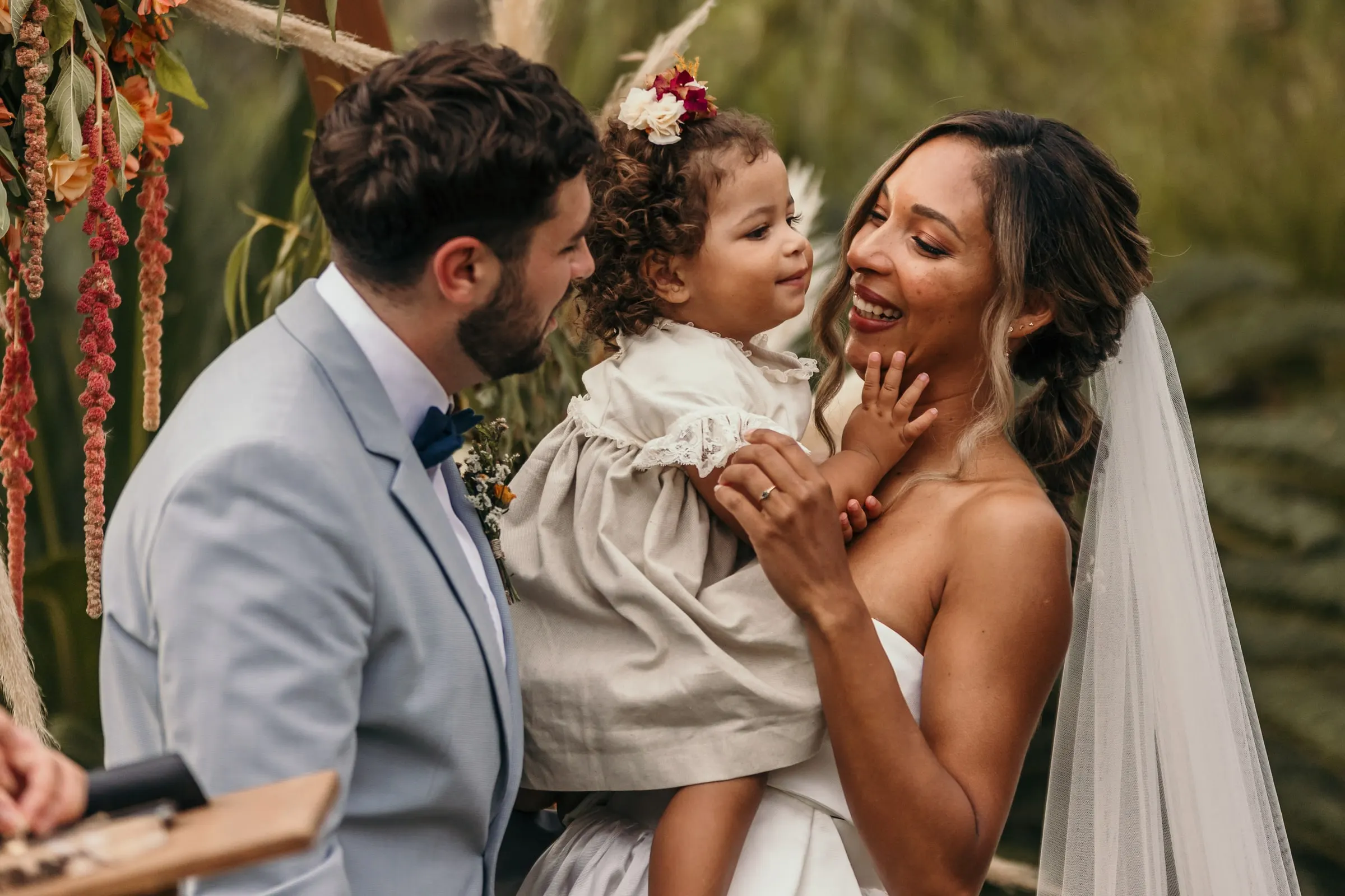 Bride and groom laughing with the little girl in her flower crown at the floral arch