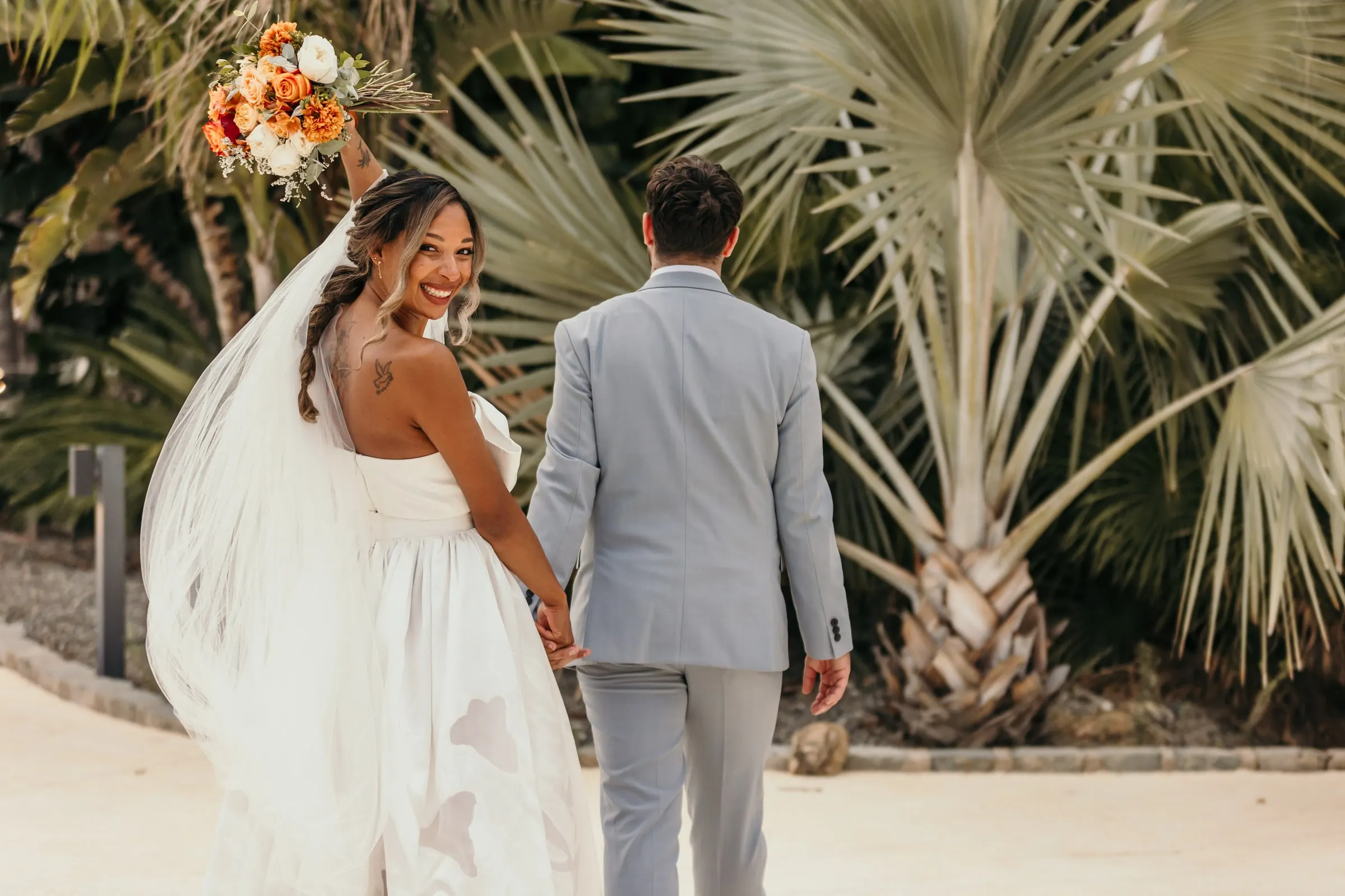 Bride looking back over her shoulder holding hands with the groom among fan palms