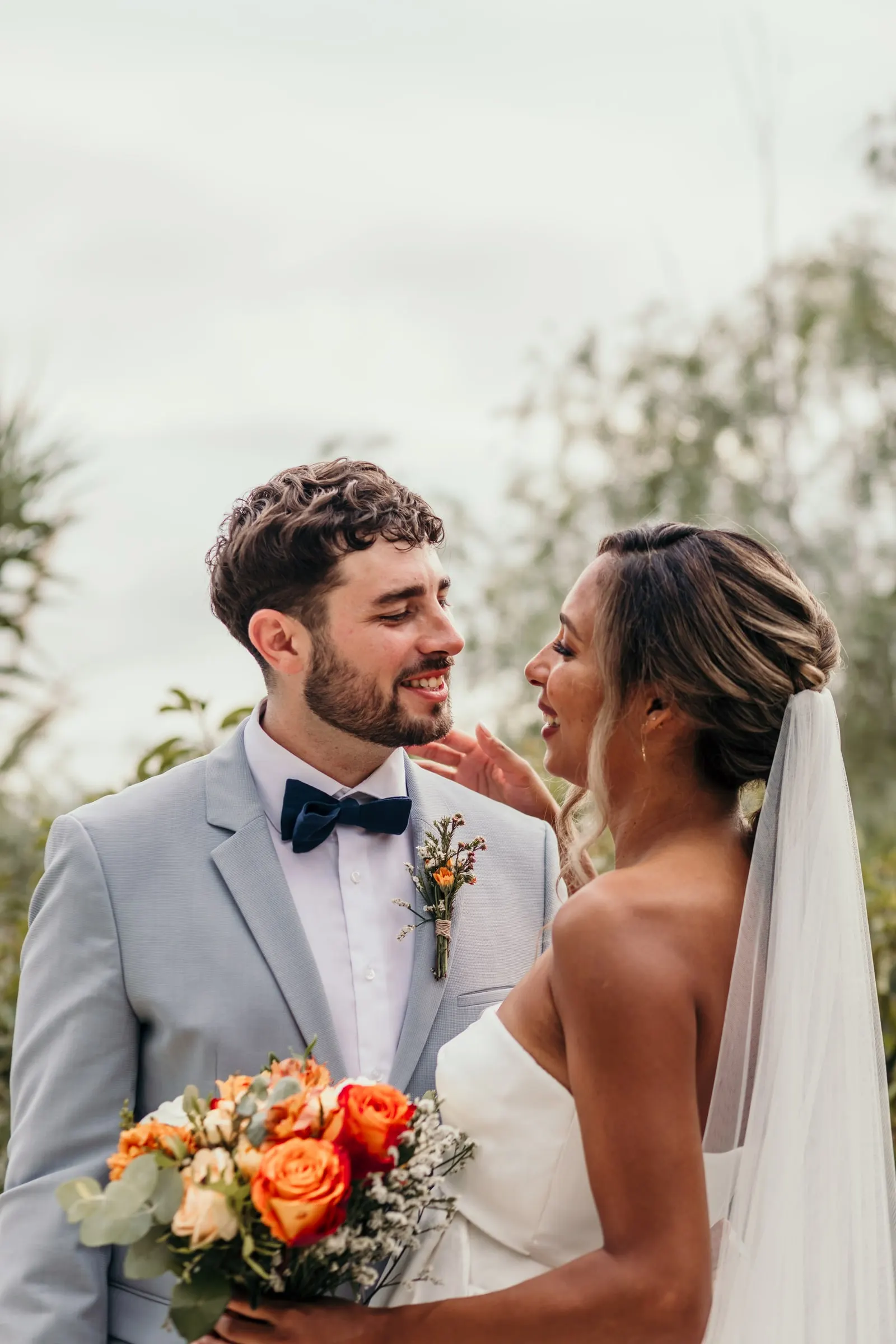 Bride touching the groom's face as they laugh, orange roses in her hand