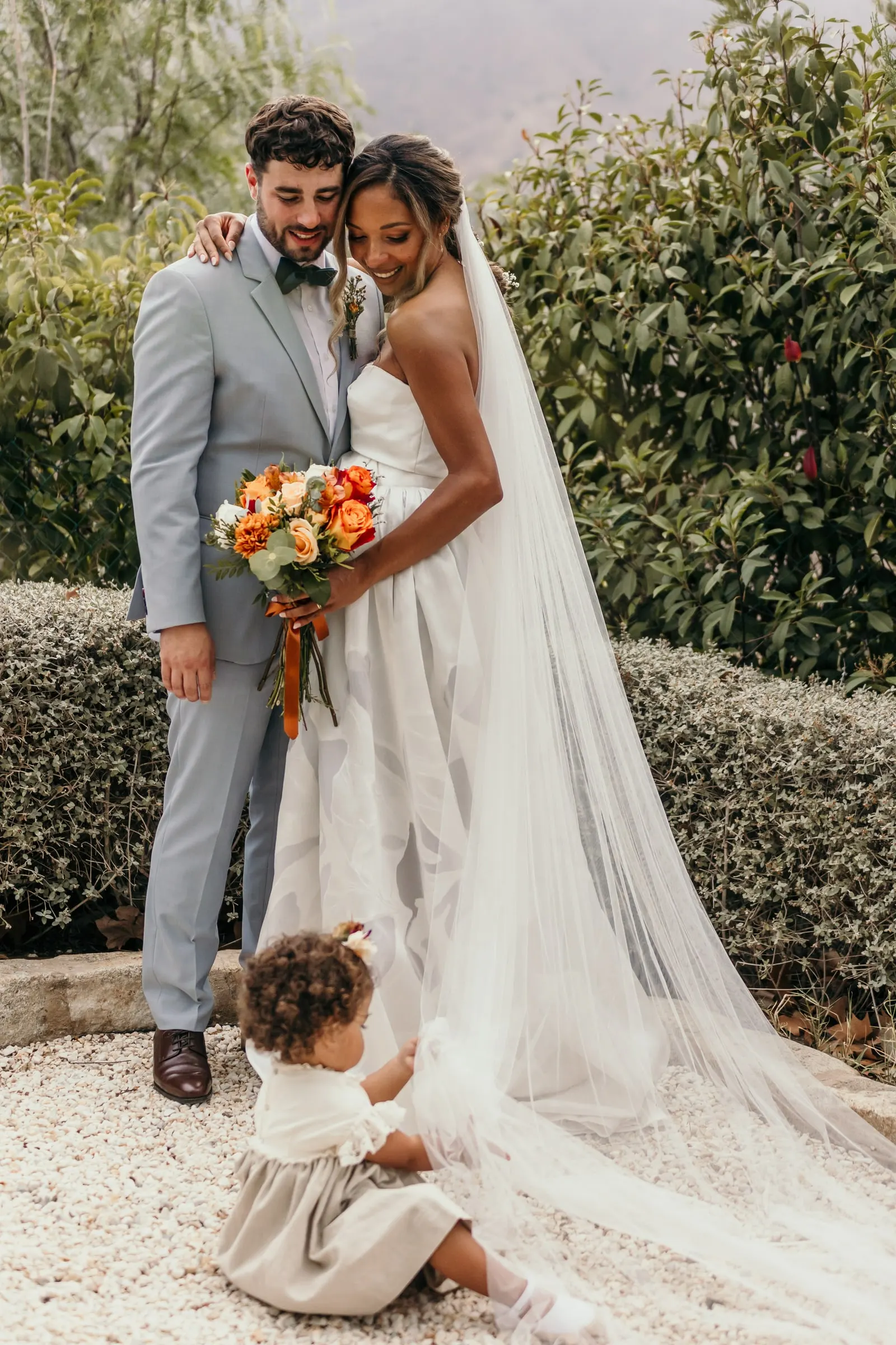 Couple portrait with the little girl playing with the bride's train on the gravel