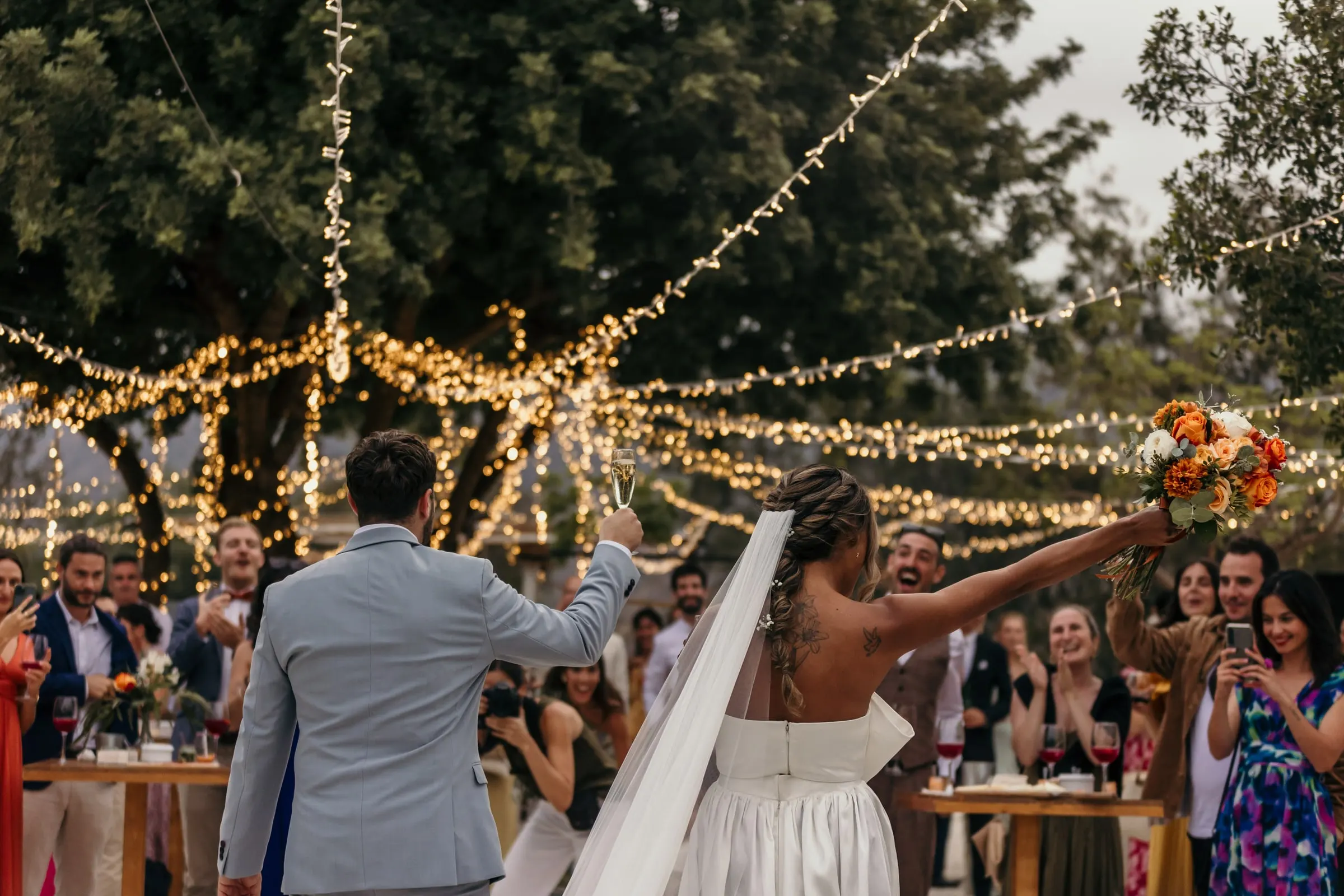 Bride raising her bouquet to the guests under strings of fairy lights