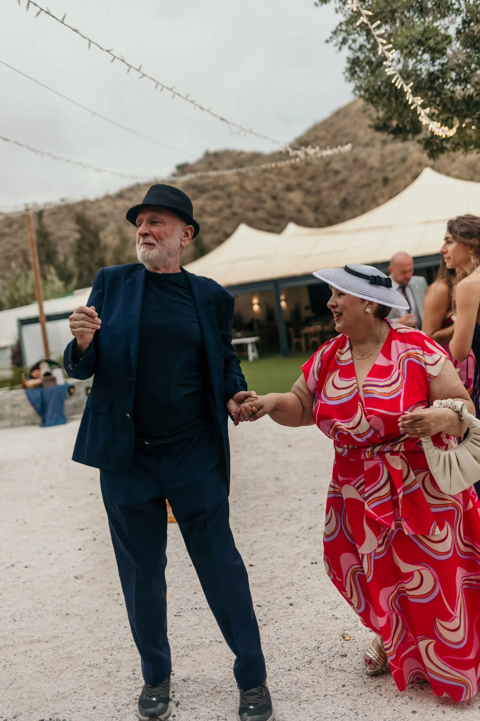 Man in the black hat dancing with a guest in a red dress and boater hat before a white marquee and a dry hillside