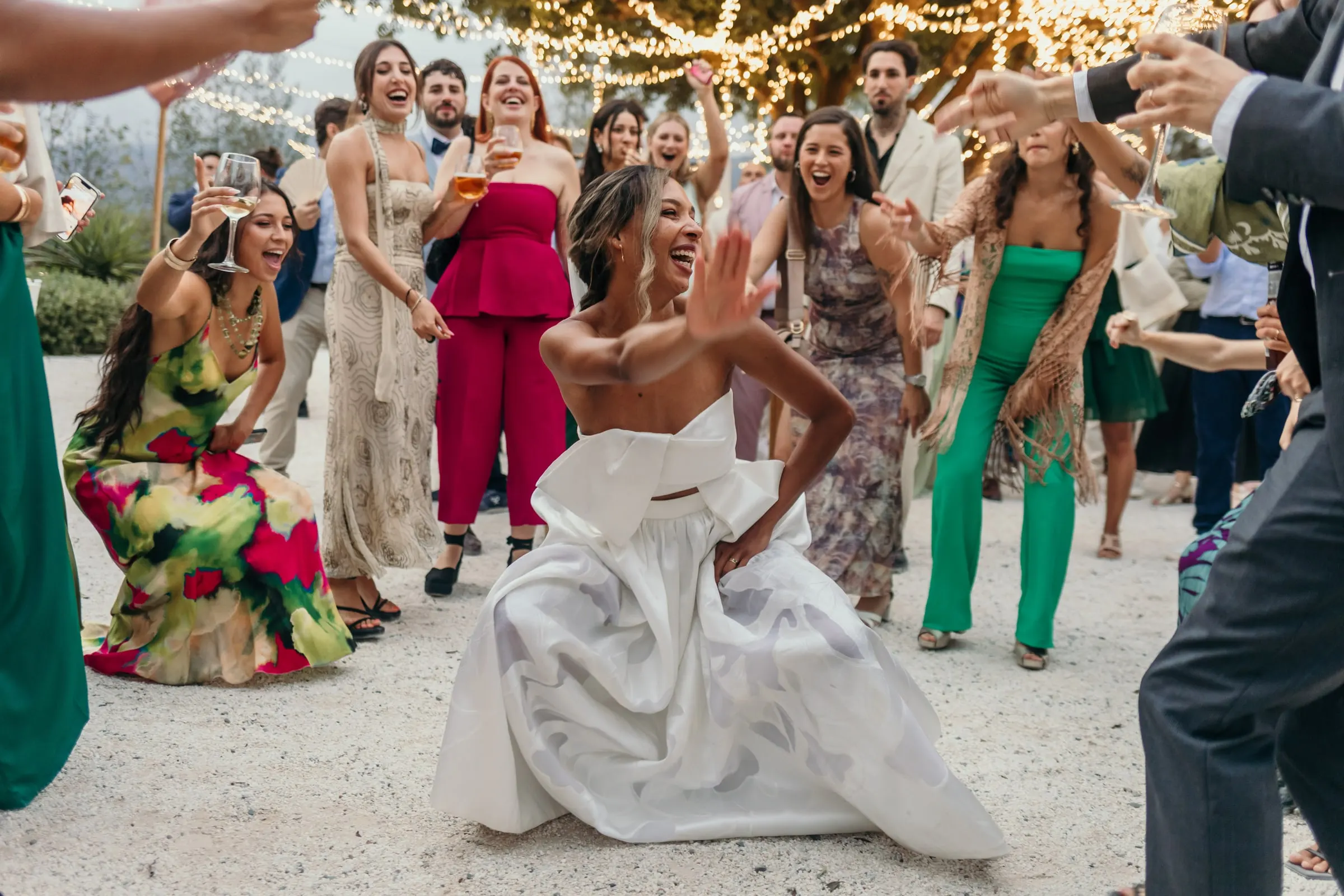 Bride crouching to dance among guests in colourful outfits on the gravel