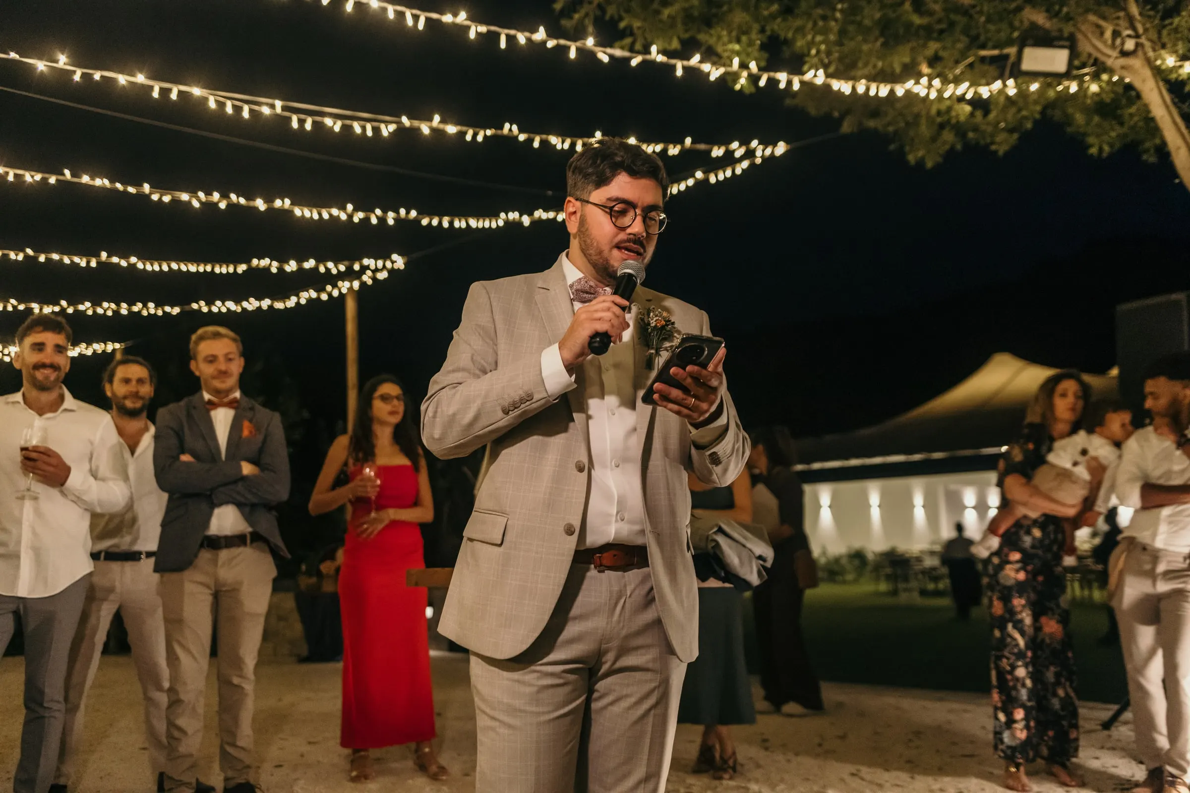 Groomsman in a beige suit speaking into the microphone at the outdoor dinner
