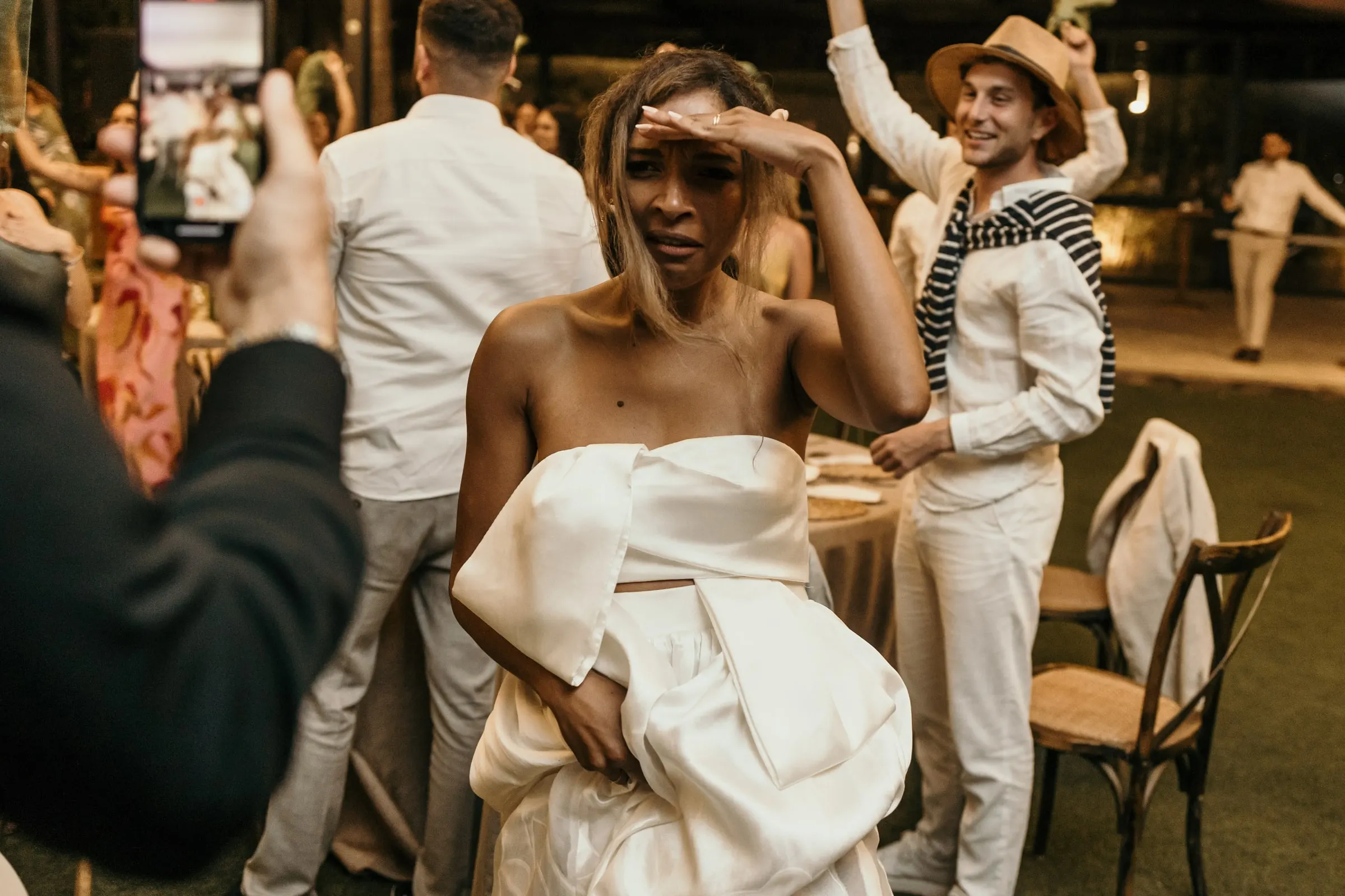 Bride shielding her eyes on the dance floor as a guest in a straw hat cheers