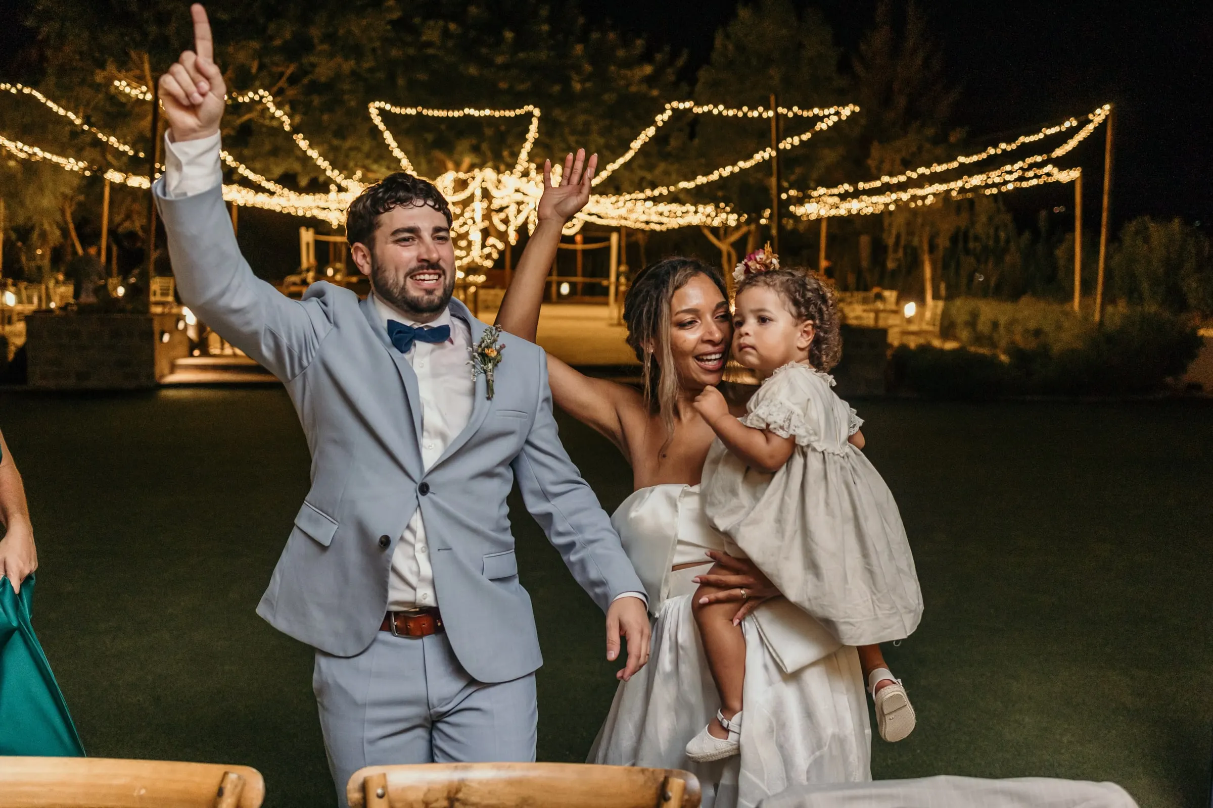Groom raising his arm as the bride holds the little girl, fairy lights behind