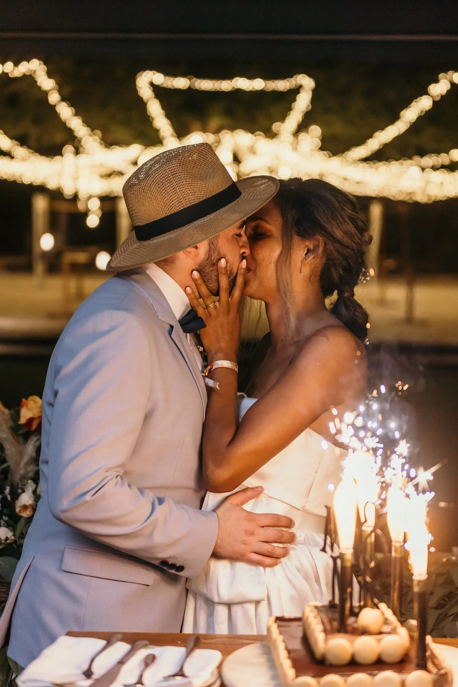 Groom in a straw hat kissing the bride beside the sparklers on the cake