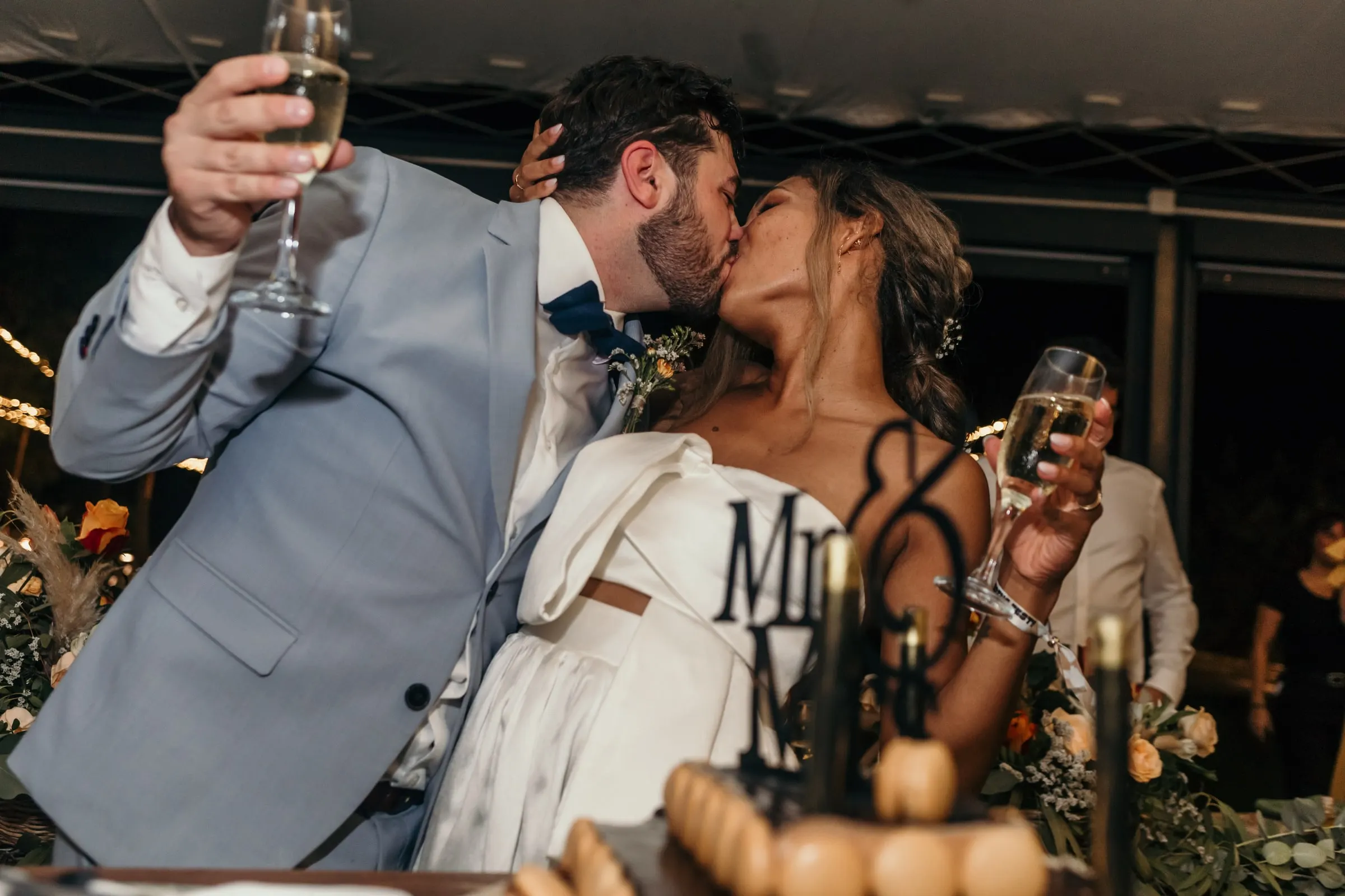 Couple kissing with glasses of cava raised by the cake table