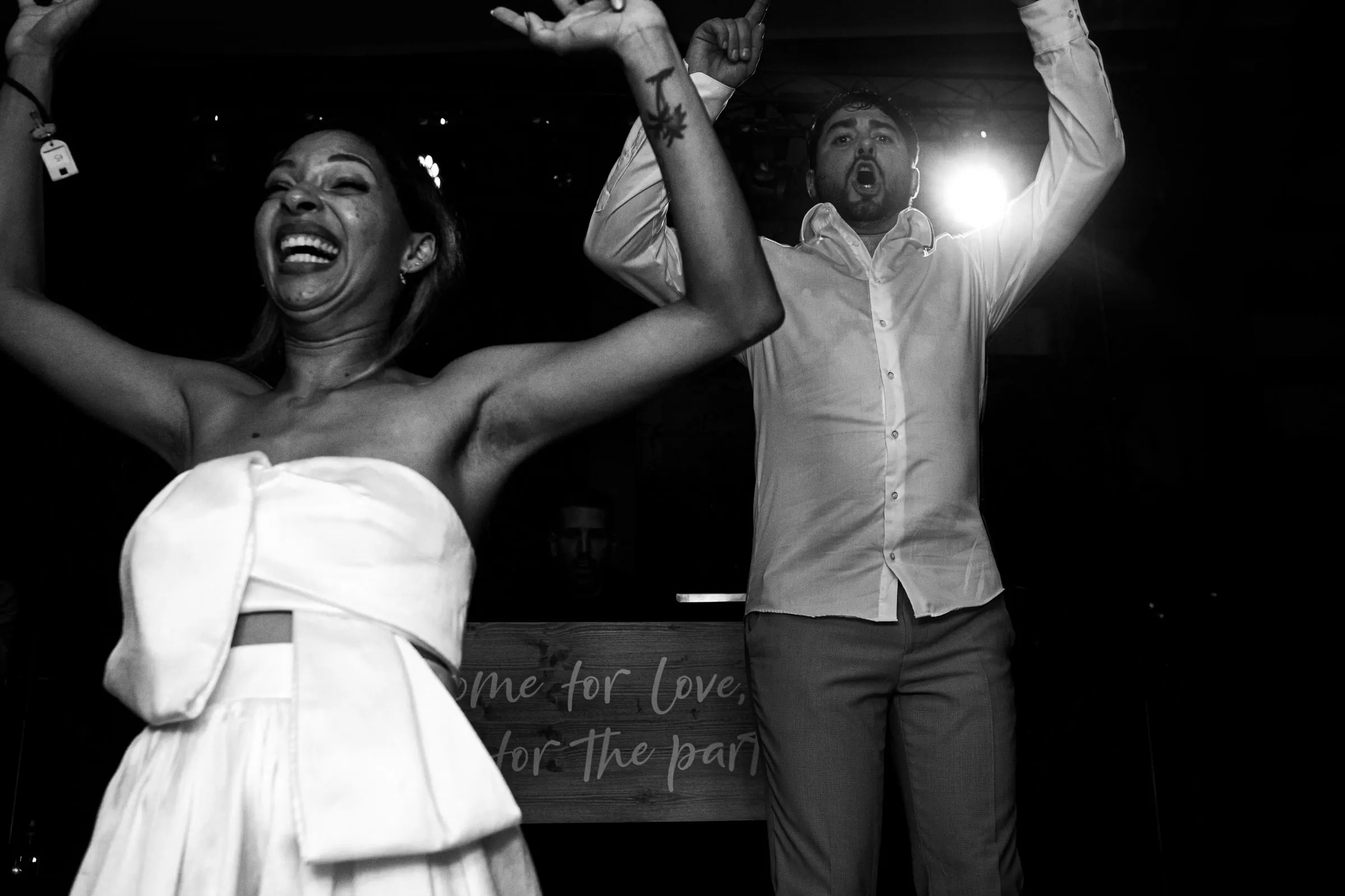 Black and white photo of the bride and groom with their arms up in front of a 'come for love, stay for the party' sign