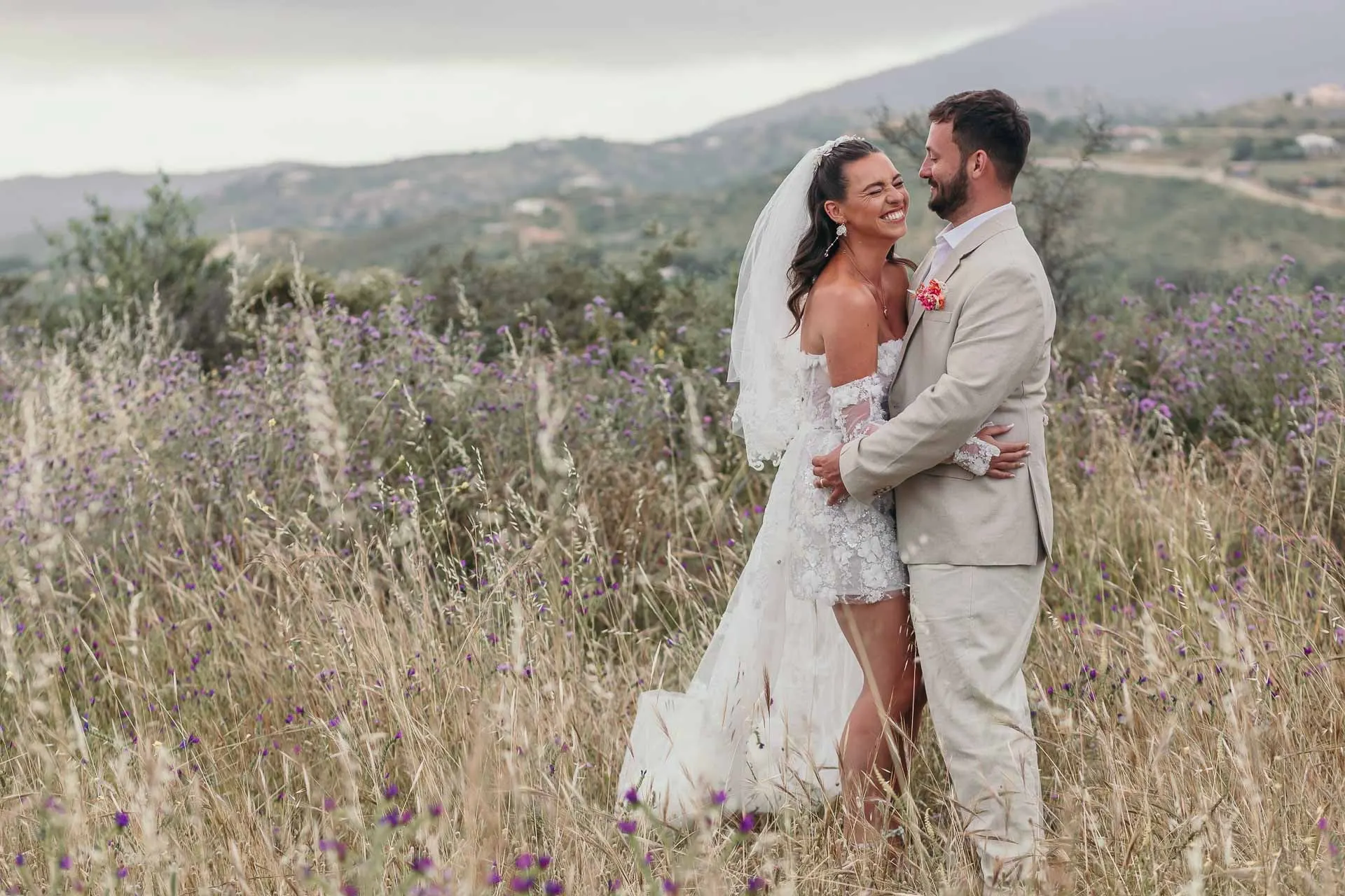 Bride and groom embracing among wild grasses and purple flowers, hills behind, Hacienda San José, Mijas