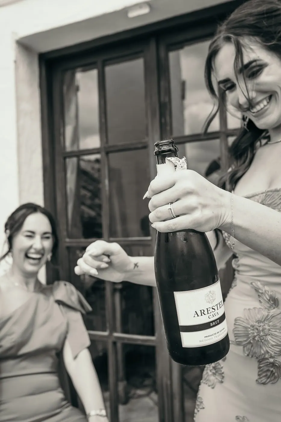 Black and white of two bridesmaids laughing as one opens a bottle of cava by a window, Hacienda San José, Mijas