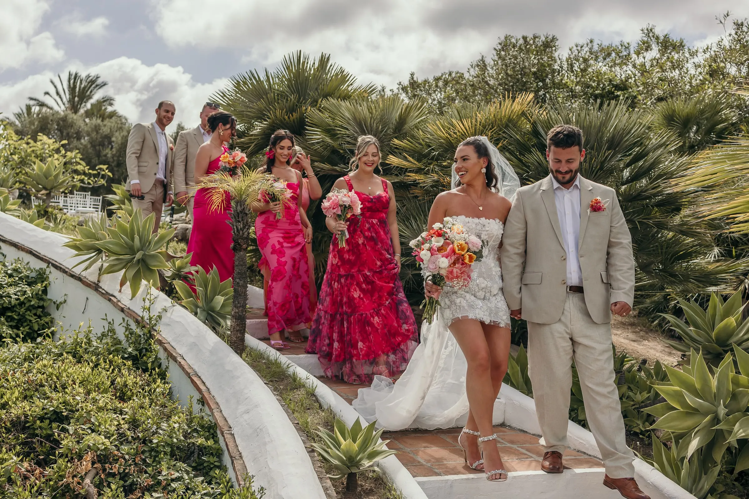 Bride and groom lead the bridesmaids in pink down a curved garden path among palms, Hacienda San José, Mijas