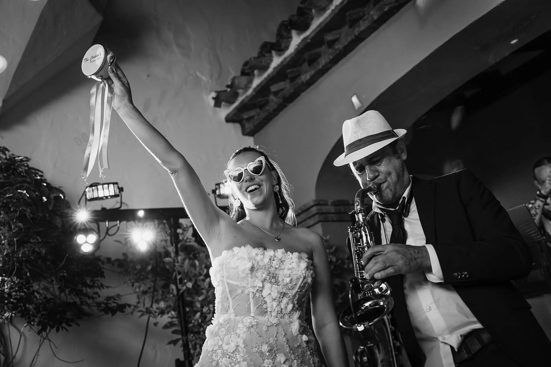 Black and white of the bride in heart sunglasses dancing beside the saxophonist, arm raised, Hacienda San José, Mijas
