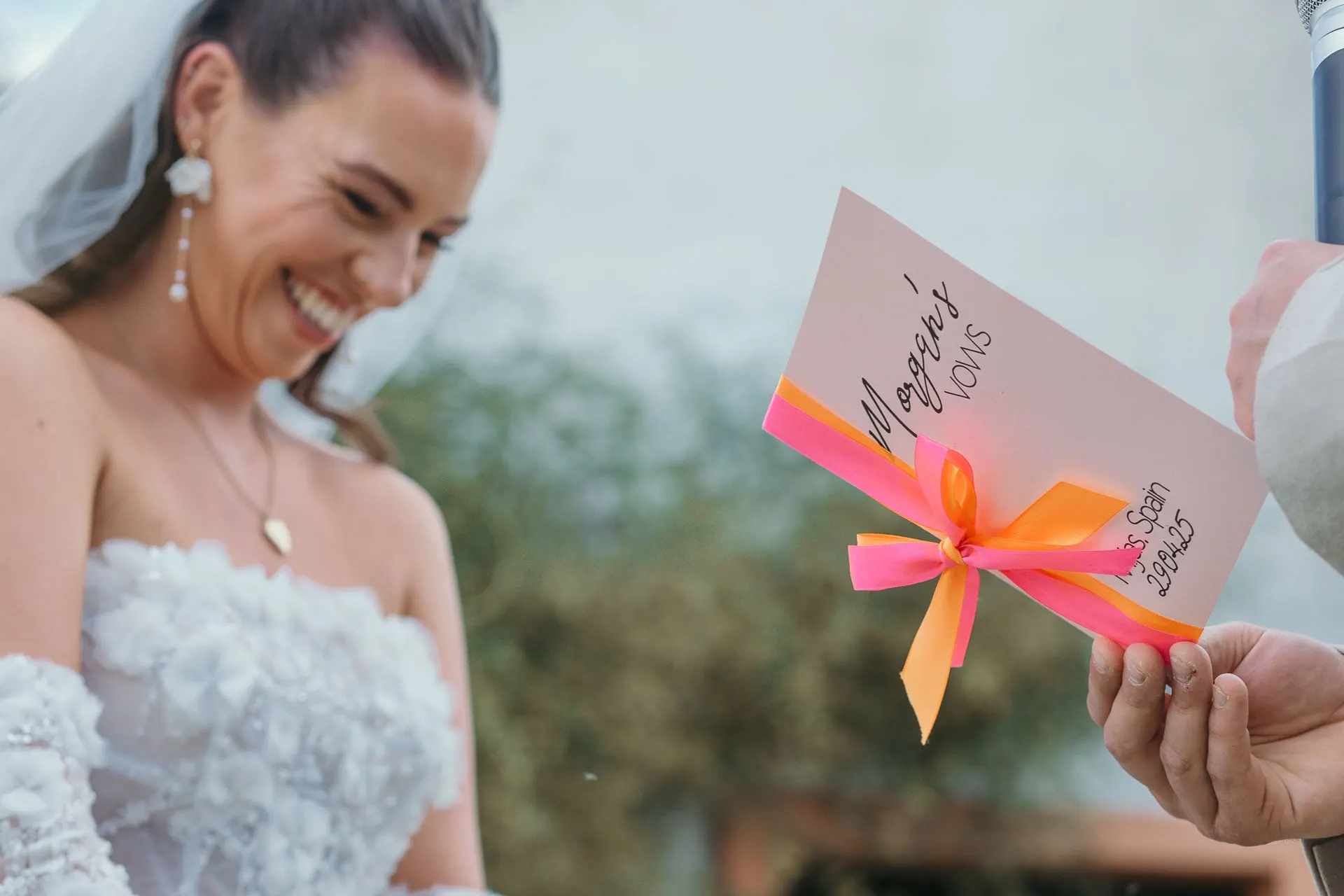 Bride laughing as a hand holds out the groom's vows card tied with pink ribbon, Hacienda San José, Mijas