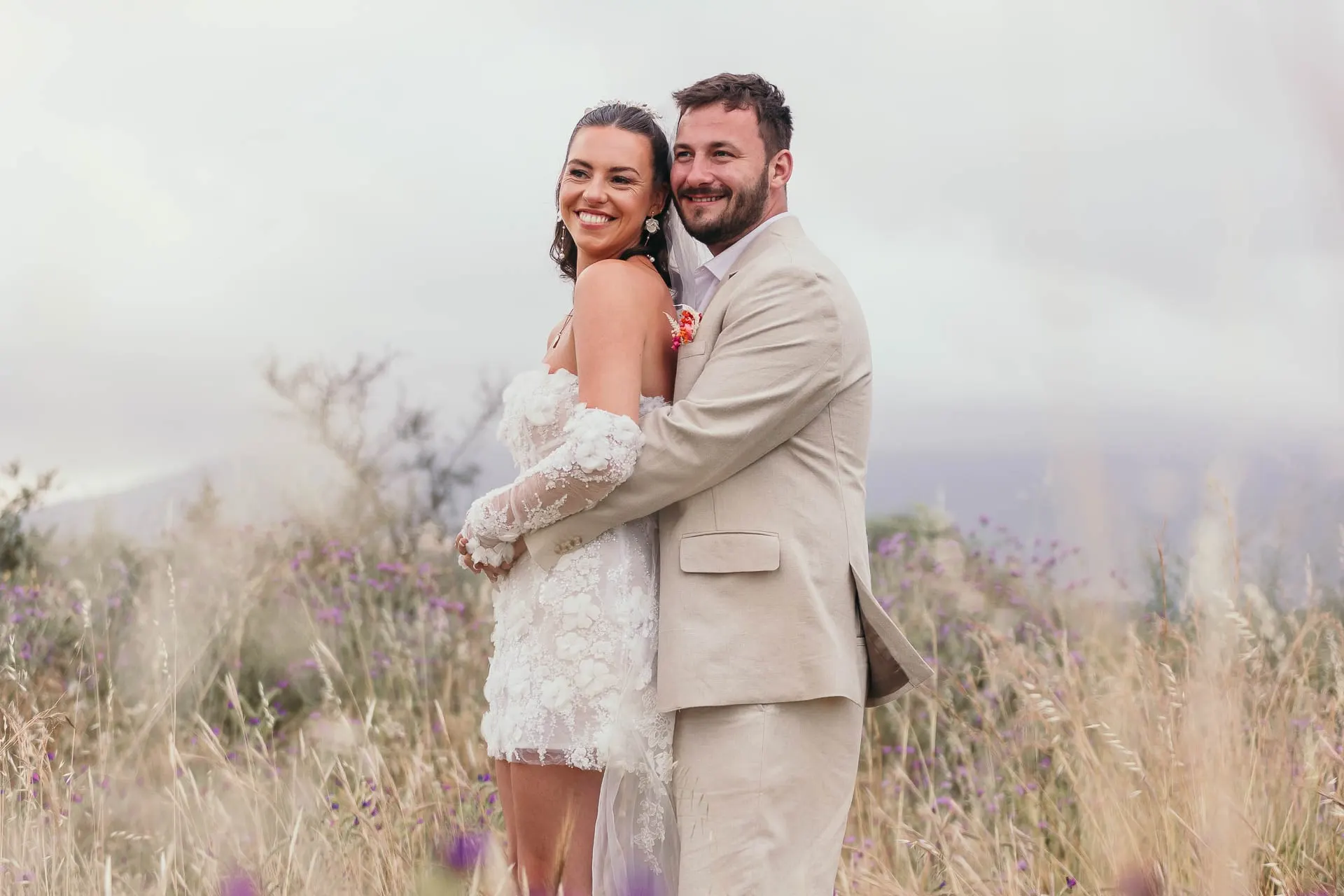 Groom hugging the bride from behind as they smile at the camera in a wild meadow, Hacienda San José, Mijas