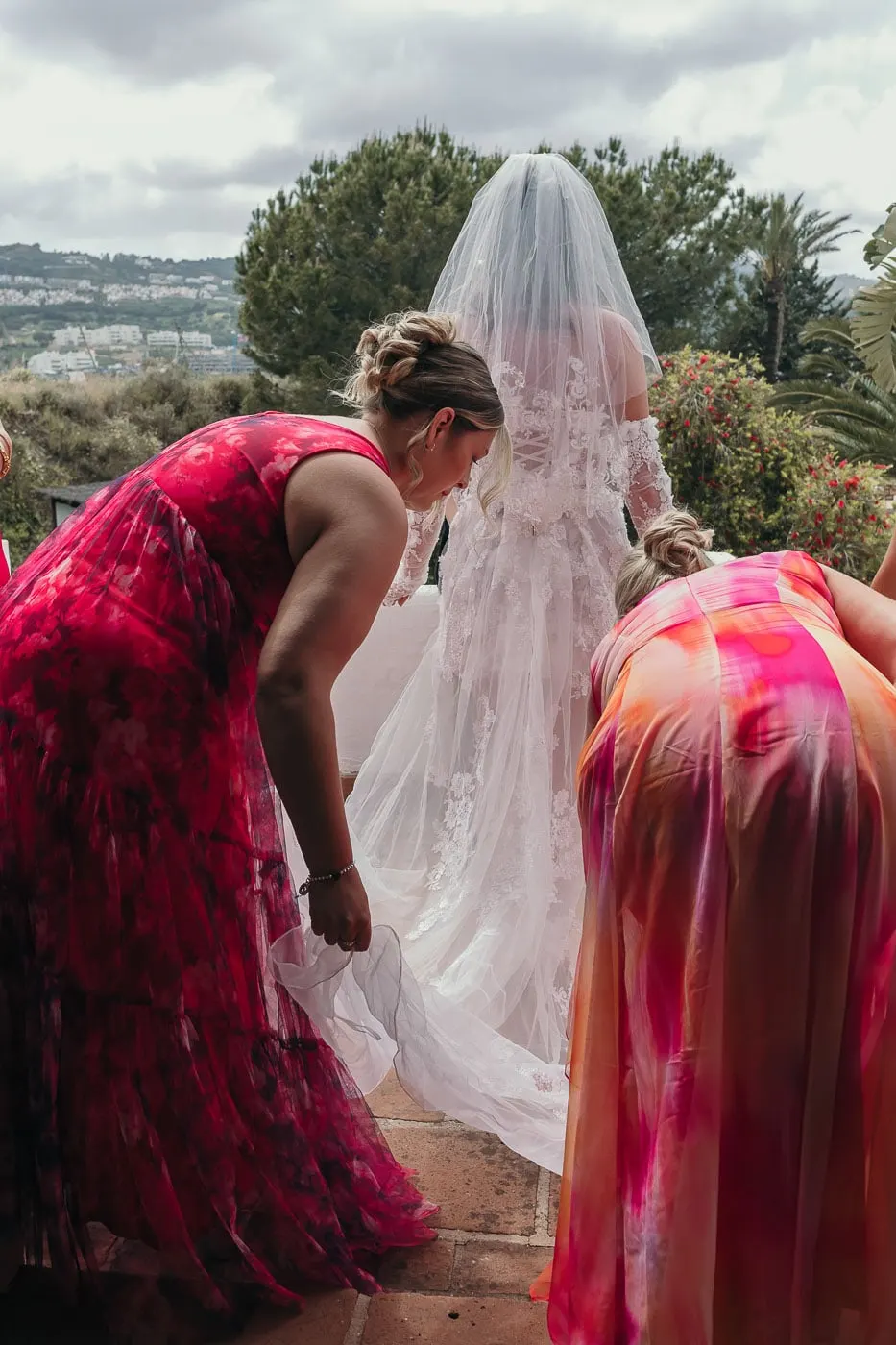 Two bridesmaids in pink straighten the bride's long veil and train on the steps, Hacienda San José, Mijas