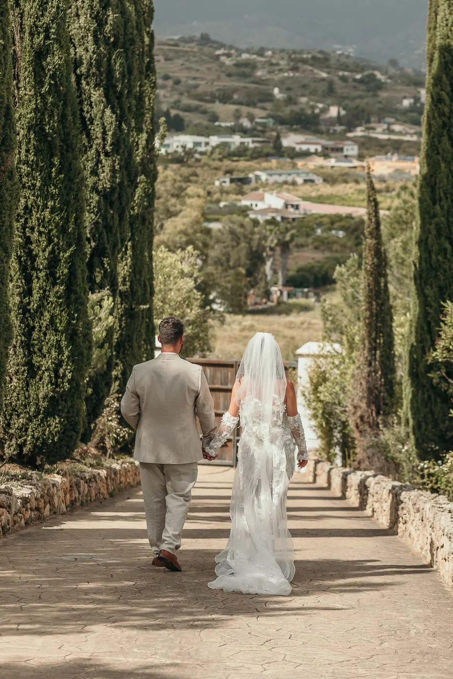 Bride and groom walking hand in hand down a cypress-lined avenue towards the valley, Hacienda San José, Mijas