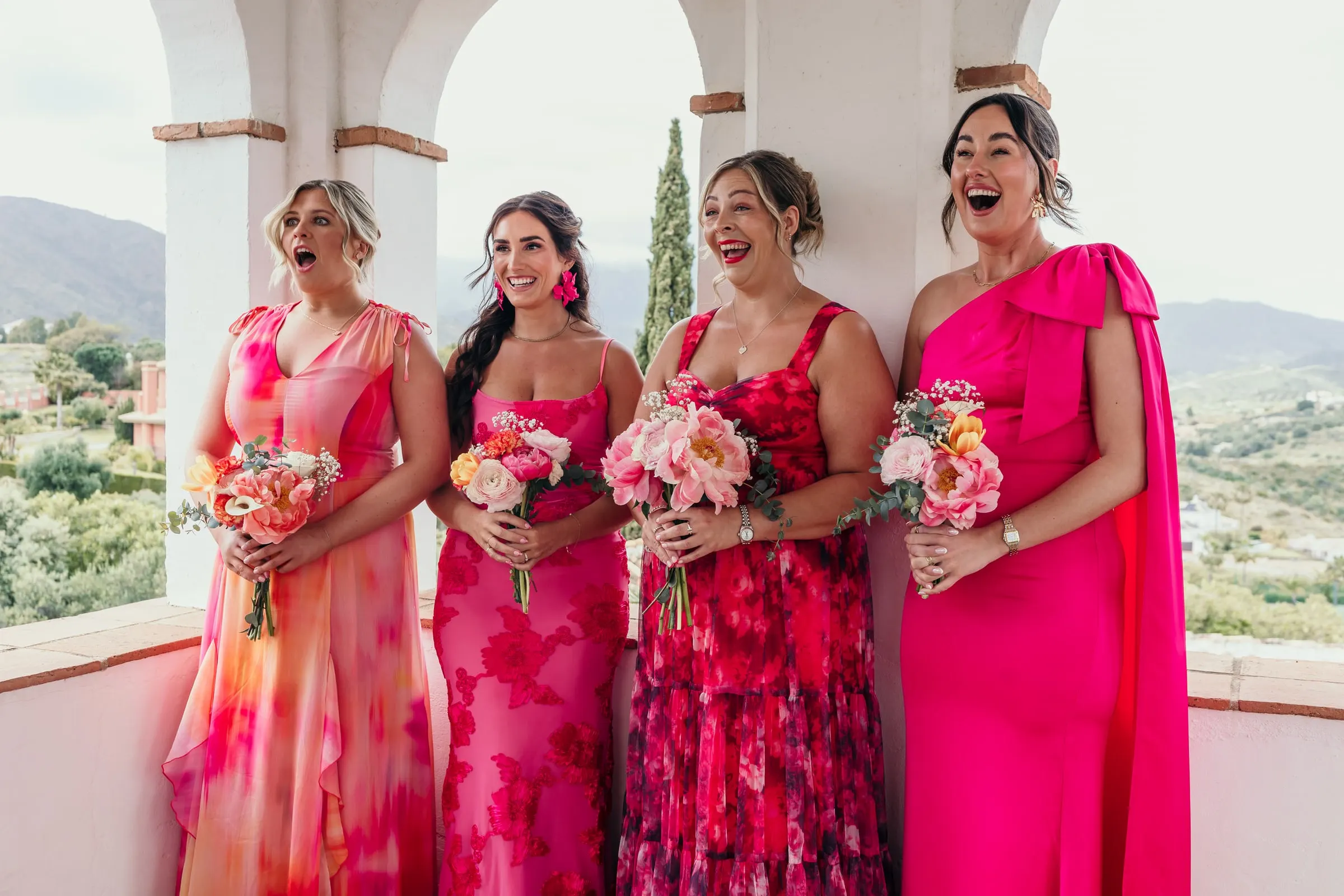 Four bridesmaids in pink dresses gasp and laugh in an arched loggia, bouquets in hand, Hacienda San José, Mijas